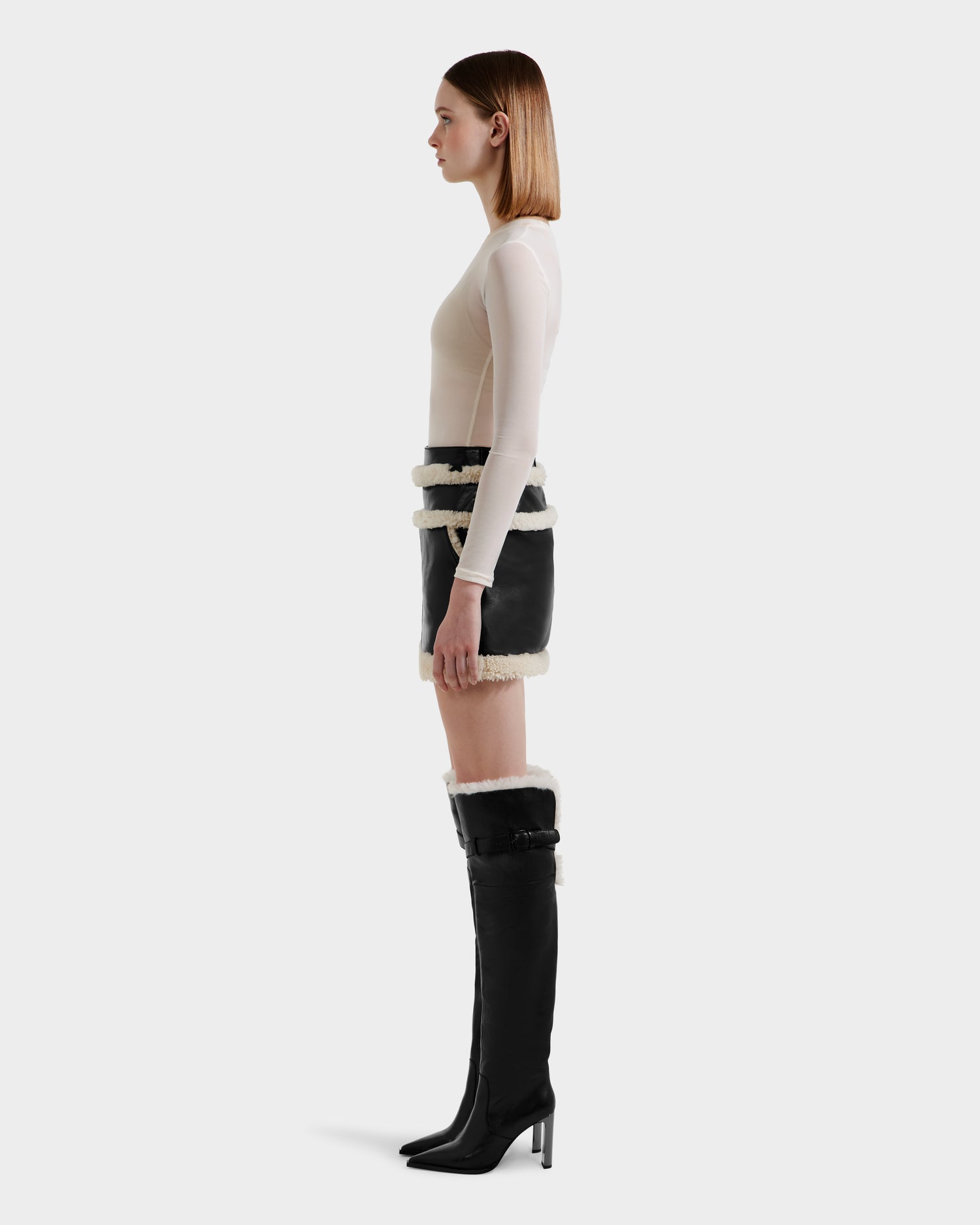 A woman in profile wears the Rudsak HAILEY - 8525609 Black lamb leather mini skirt with white shearling trim, paired with a fitted beige top and matching knee-high fur-trimmed boots, against a plain white background.