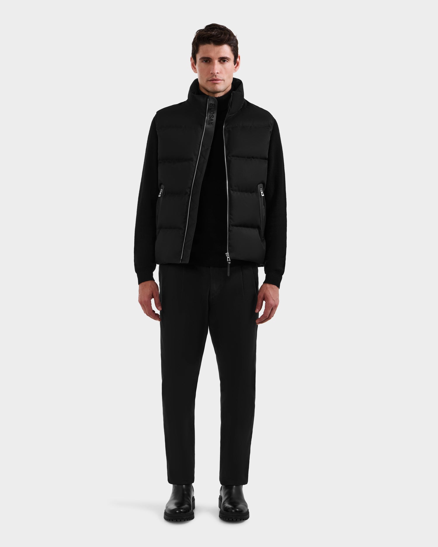 A man stands facing forward against a plain white background, wearing the Rudsak HARLOW - M125528 Black men’s puffer vest with a removable hood over a black long-sleeve shirt, black pants, and black boots.