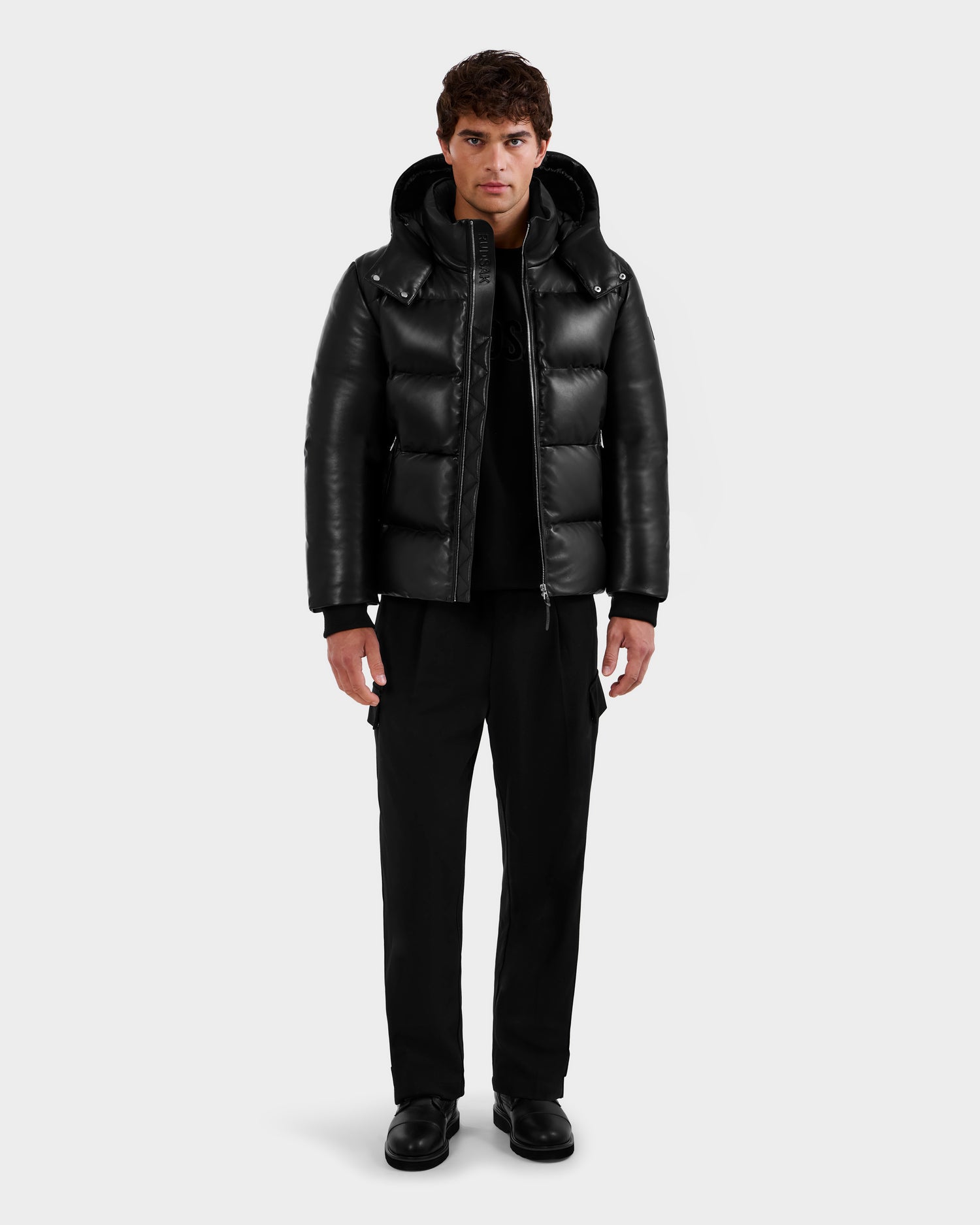 A man stands facing forward in a Rudsak HARVEY X - 6125681 Black hooded puffer jacket, paired with black pants and shoes, set against a plain light gray background.