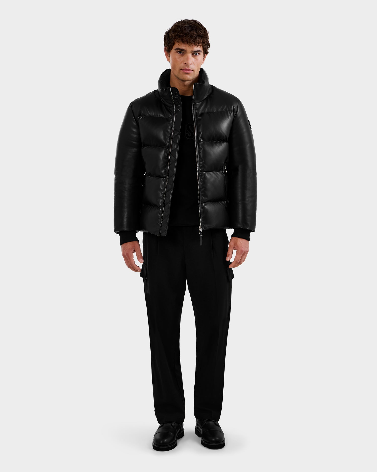 A man with short brown hair and a neutral expression stands against a plain background, wearing the Rudsak HARVEY X - 6125681 Black puffer jacket, paired with black pants and black shoes.