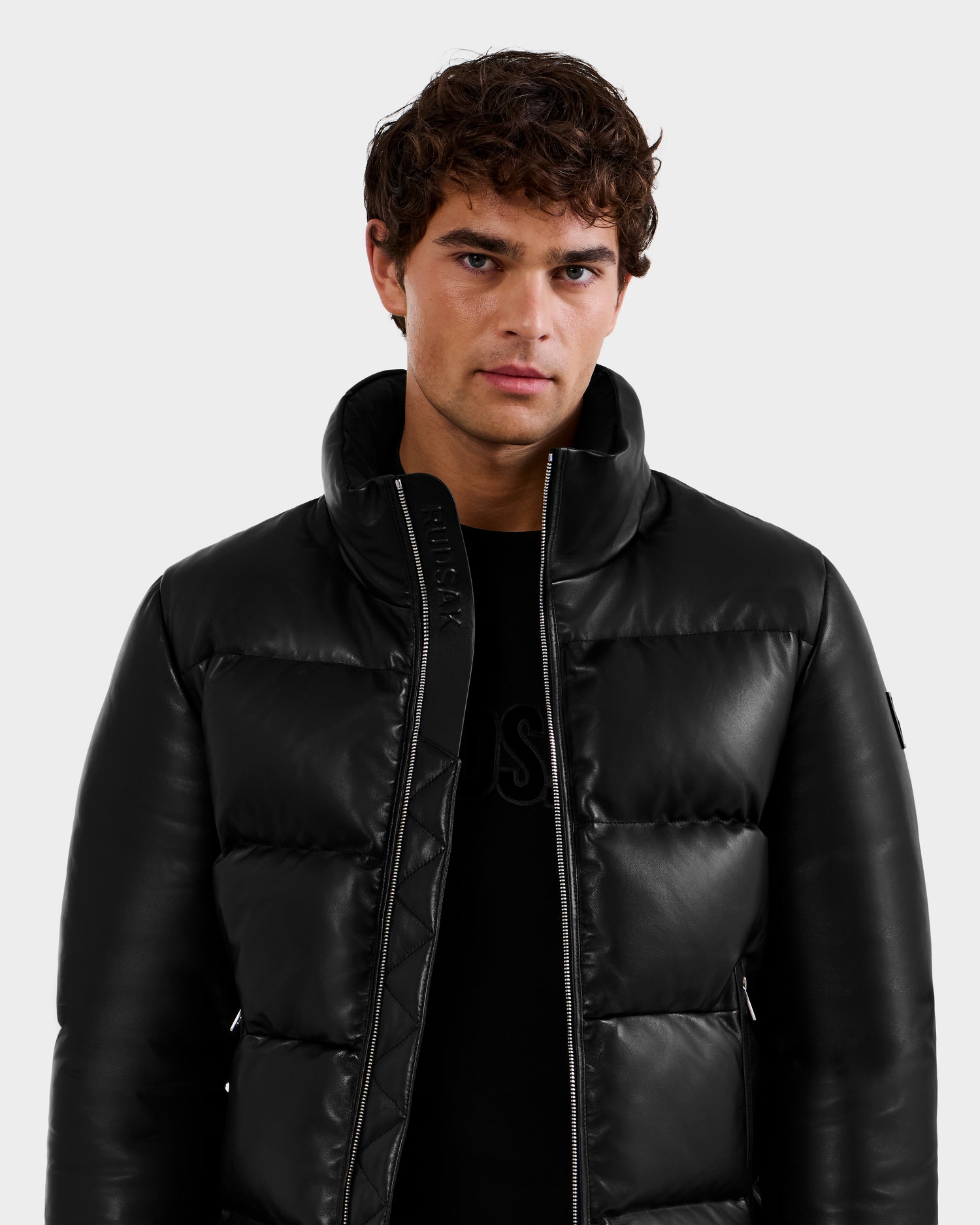 A young man with curly brown hair wears the Rudsak HARVEY X - 6125681 Black puffer jacket over a black shirt, standing against a plain light gray background and looking directly at the camera.