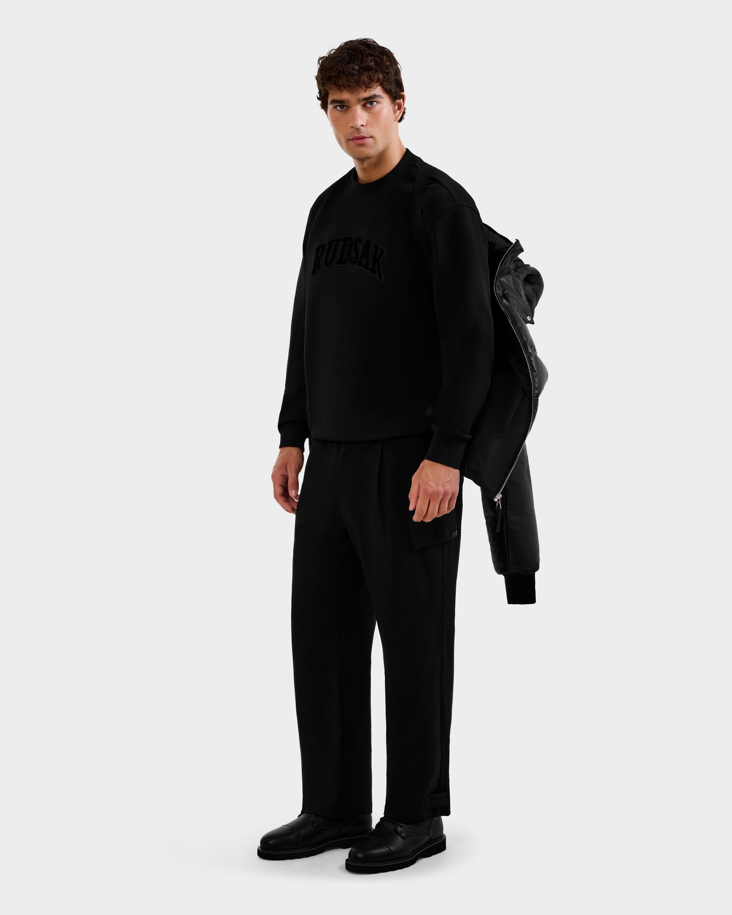 A man stands against a plain background wearing the HARVEY X - 6125681 Black jacket by Rudsak over his shoulder. Dressed in all-black, he looks directly at the camera with a neutral expression.