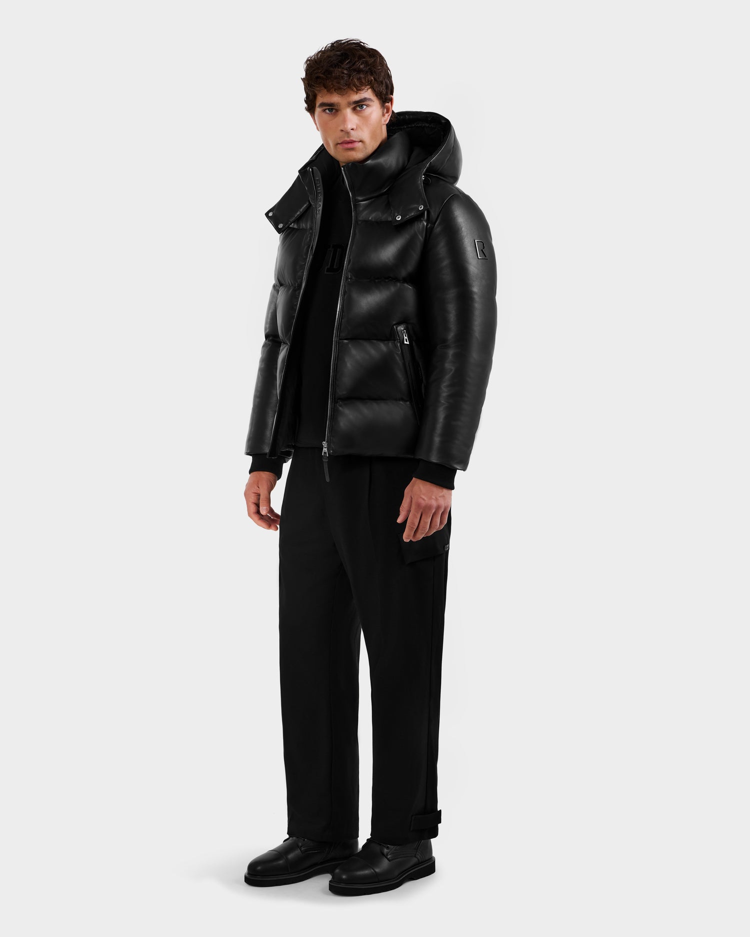 A man stands slightly turned against a light gray background, wearing the Rudsak HARVEY X - 6125681 Black puffer jacket with a high collar, paired with black pants and black shoes.