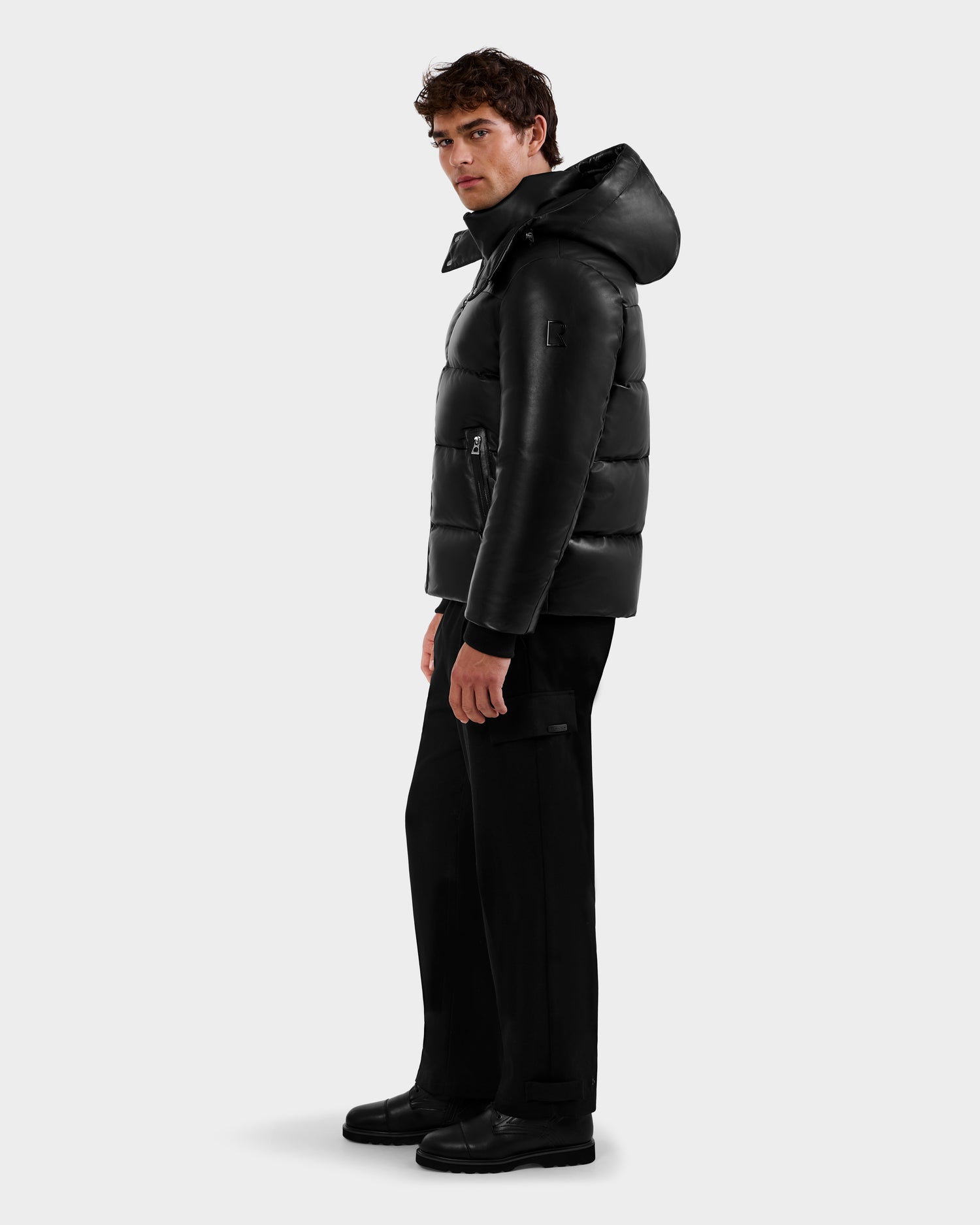 A man with curly hair, dressed in Rudsak's HARVEY X - 6125681 Black hooded puffer jacket, black pants, and black boots, stands sideways against a plain light background.
