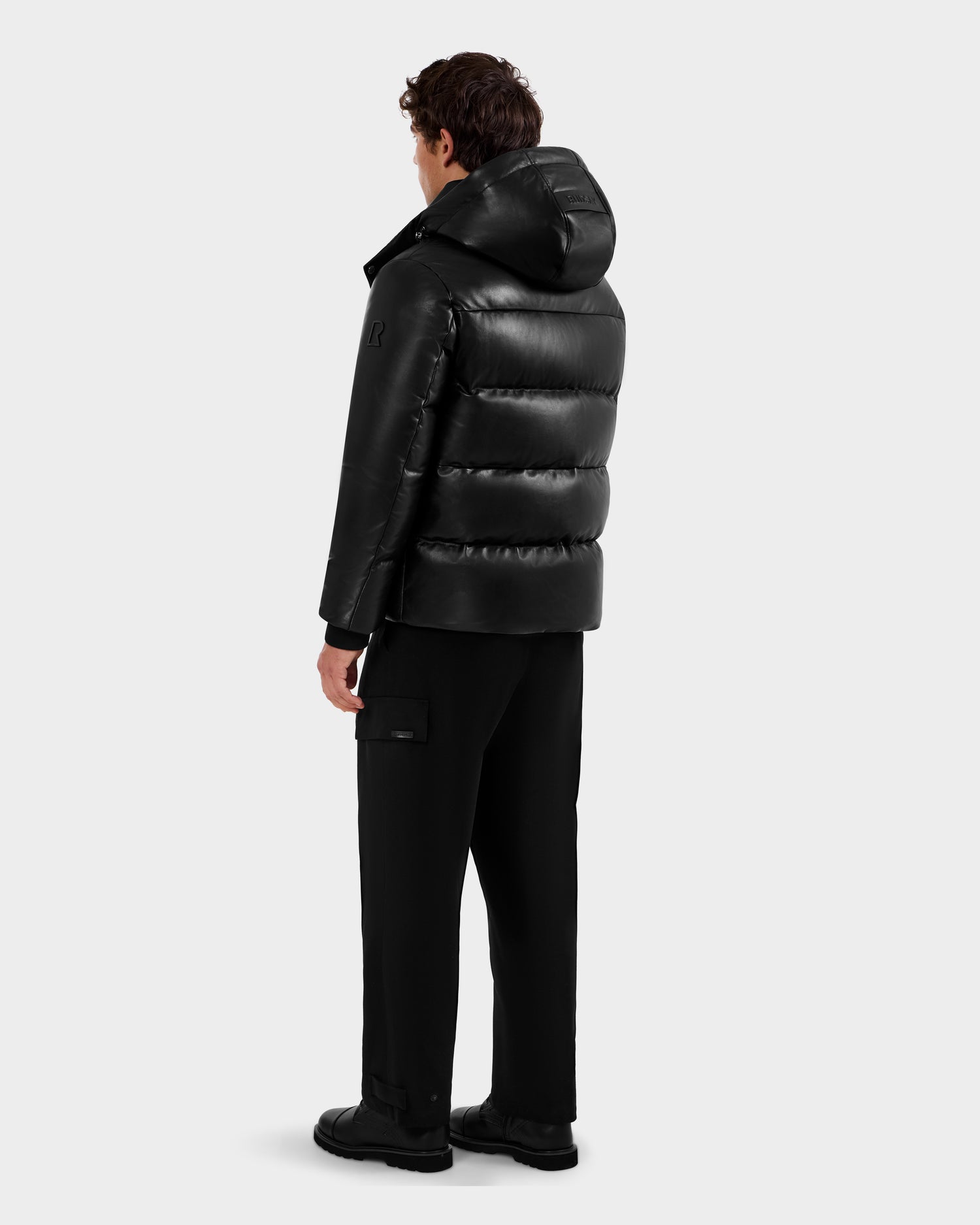 A person stands with their back to the camera, wearing the Rudsak HARVEY X - 6125681 Black puffer jacket, paired with black pants and shoes, set against a plain light gray background.