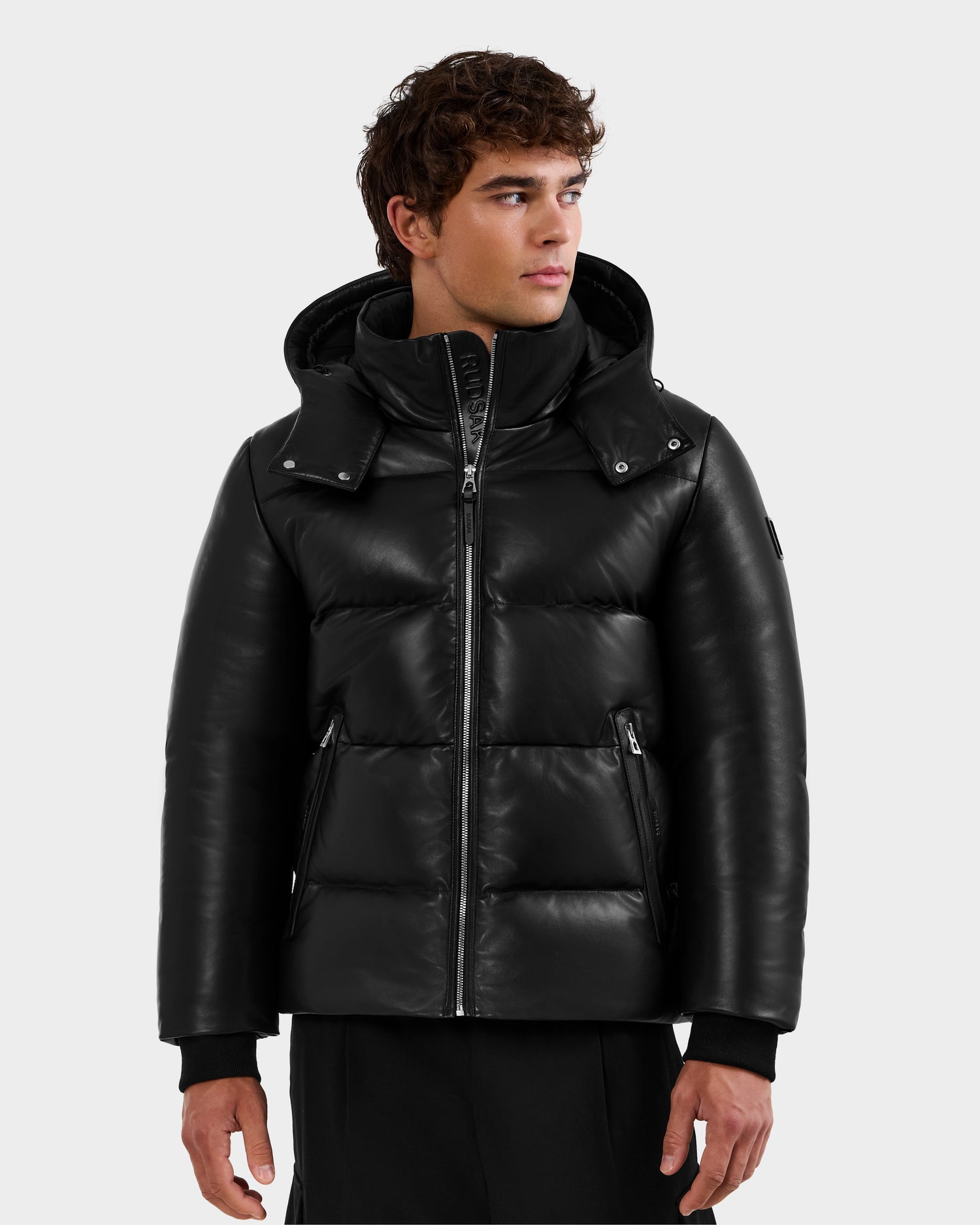 A young man in the Rudsak HARVEY X - 6125681 Black leather puffer with hood and zippered pockets stands against a plain light background, looking to his right with a neutral expression.