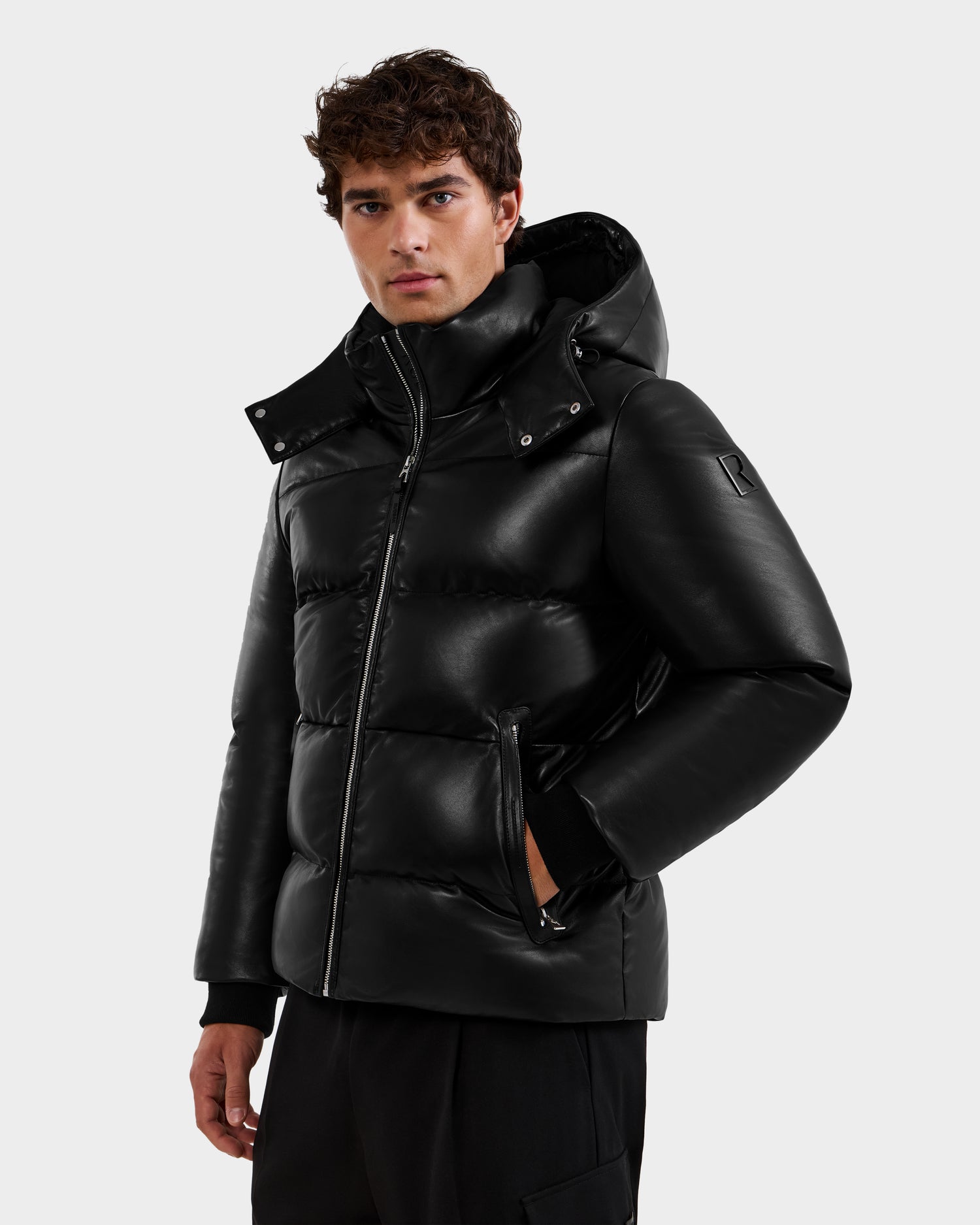 A man wears the Rudsak HARVEY X - 6125681 Black puffer jacket with a hood and zippered pockets, standing against a plain light gray background and looking at the camera with a neutral expression.