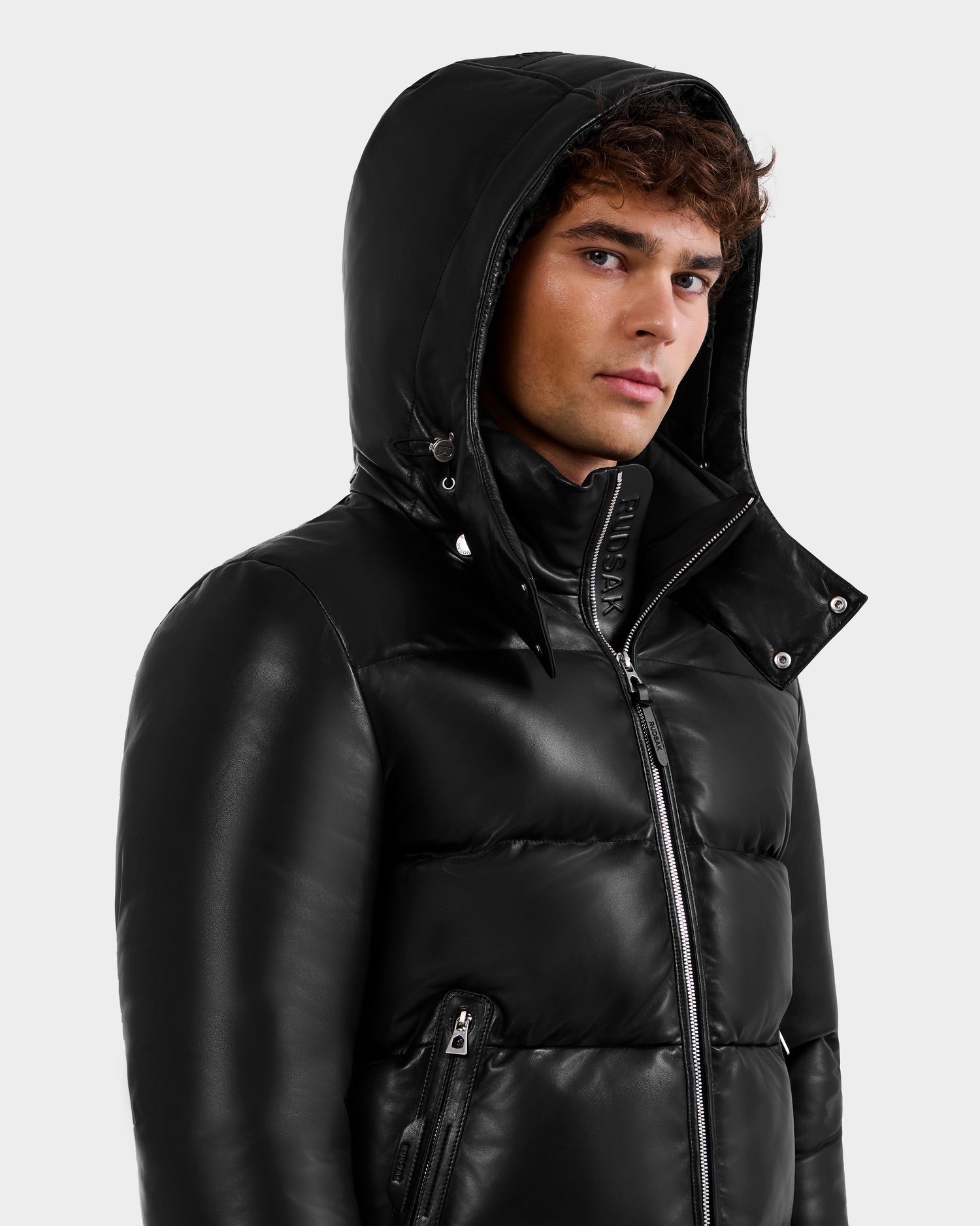 A man with curly brown hair wears the Rudsak HARVEY X - 6125681 Black puffer jacket featuring a hood, high collar, and front zipper. He gazes slightly to the side with a neutral expression against a plain light background.