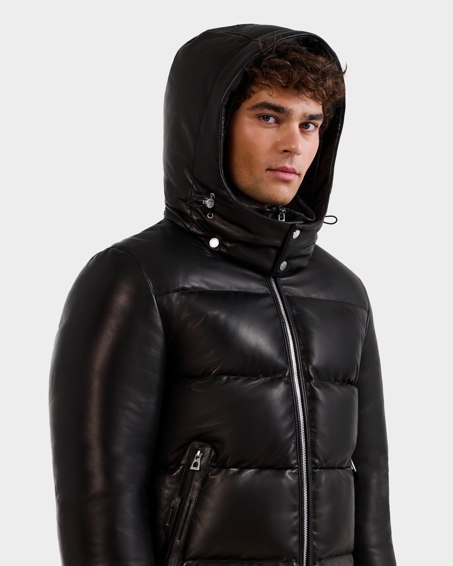 A young man with curly hair models the Rudsak HARVEY X - 6125681 Black shiny hooded puffer jacket, looking slightly to the side against a plain white background.