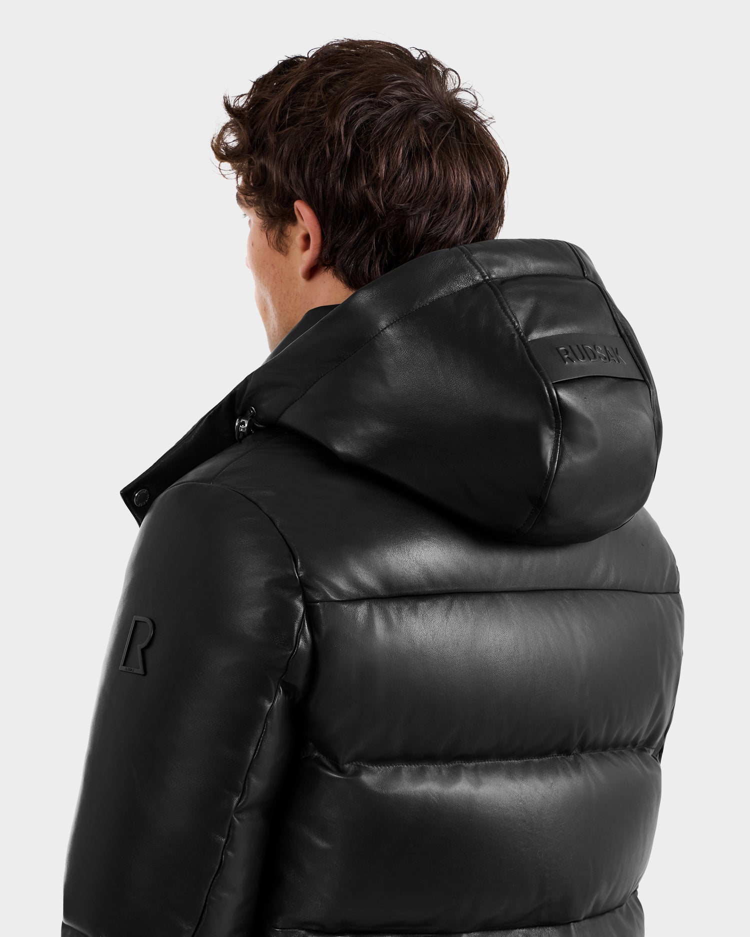 A person with short curly hair is seen from behind, wearing the Rudsak HARVEY X - 6125681 Black puffer jacket with a shiny finish and embossed text on the hood, against a plain background.