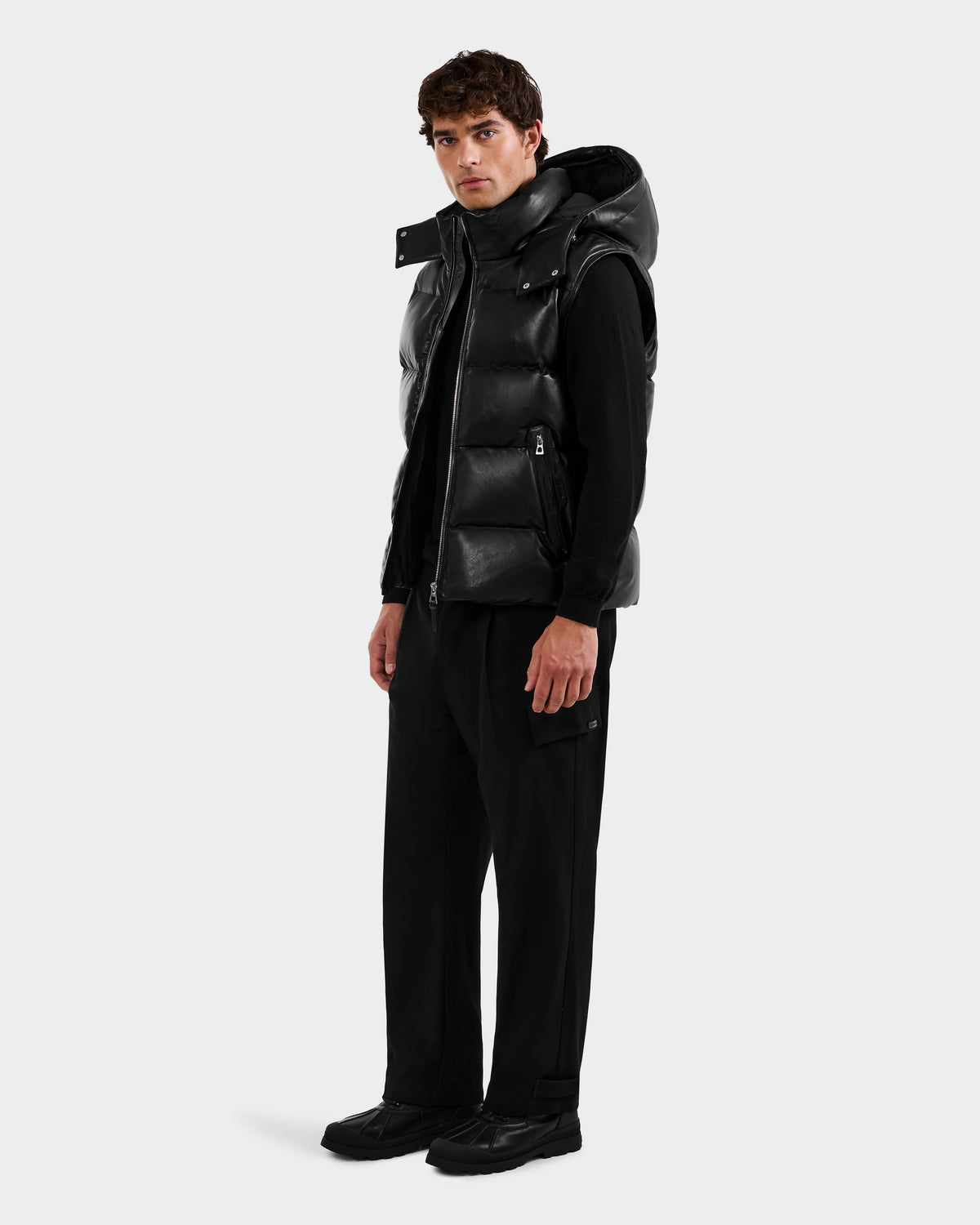 A man faces slightly left, wearing the HENRY X - 6125679 Black puffer vest by Rudsak over a black hoodie, with black trousers and shoes, set against a plain light gray background.