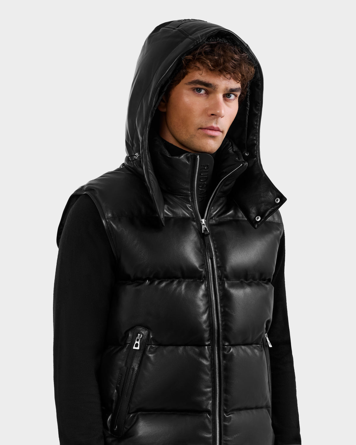 A young man with curly hair wears the Rudsak HENRY X - 6125679 Black puffer vest with a hood over a black long-sleeve shirt, standing against a plain light background and looking slightly to the side.