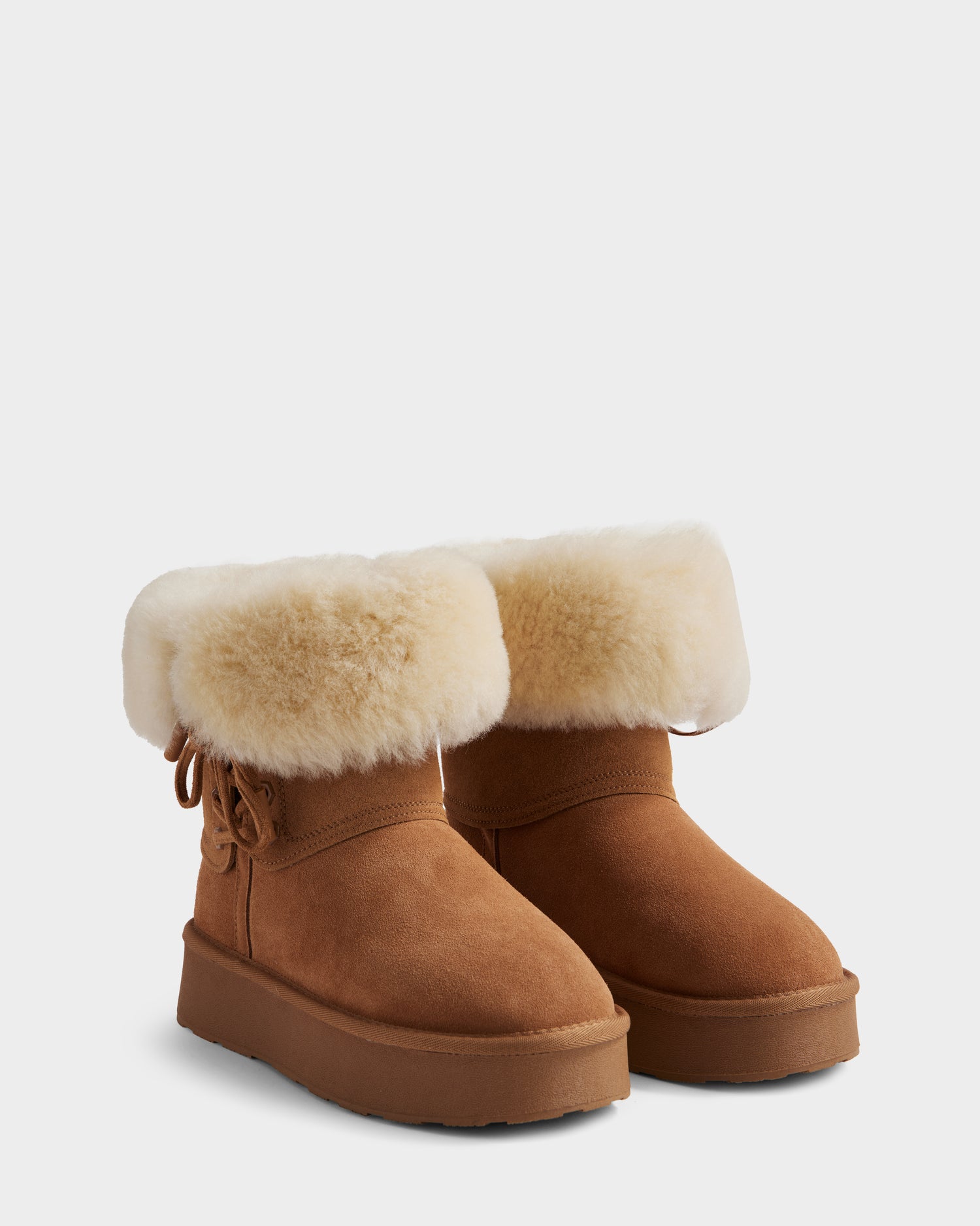 The Rudsak INESA - L225026 Tan boots feature brown suede, a fluffy white wool lining at the top, side bow detail, and flat rubber soles, shown against a plain light gray background.