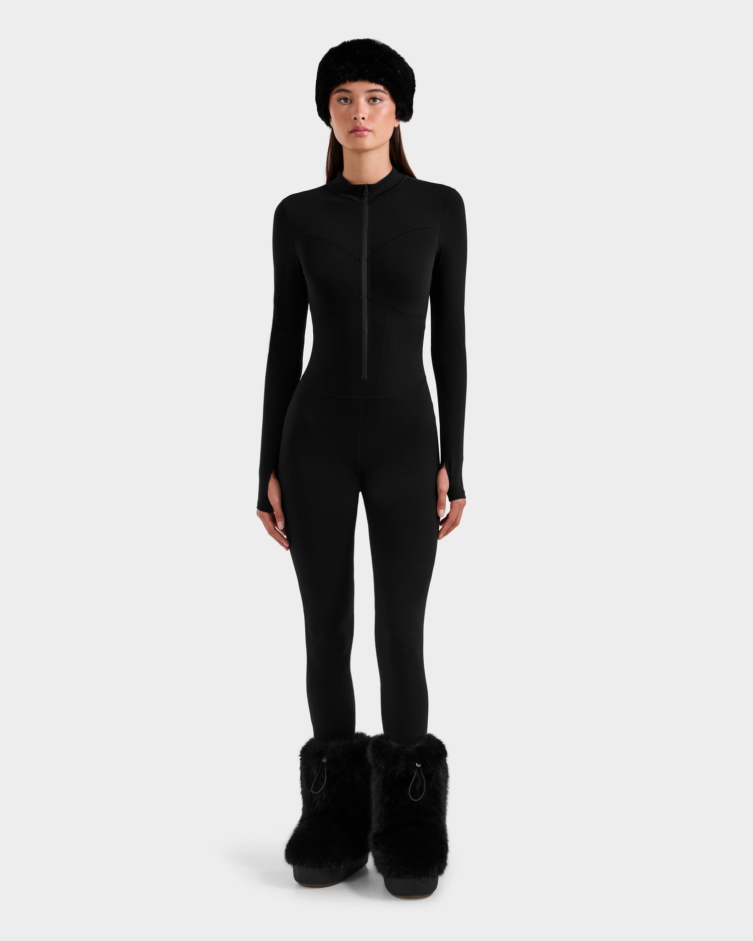 A woman in the Rudsak IVY - 8524599 Black stretch bodysuit, paired with a black fur hat and oversized fur boots, channels ski-chic style against a plain white background.