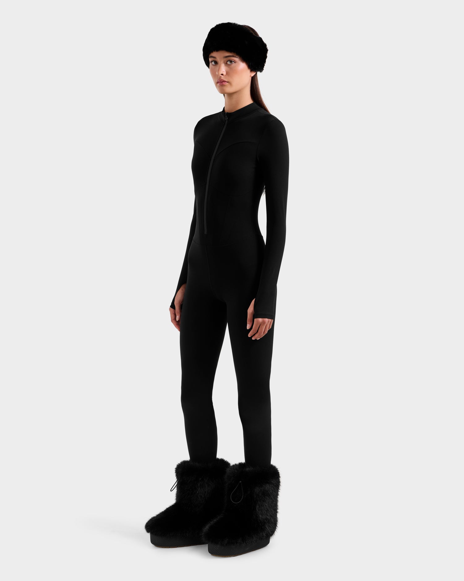 A woman in ski-chic style poses against a plain white background, wearing the Rudsak IVY - 8524599 Black fitted women's stretch bodysuit, paired with a black furry hat and oversized black furry boots.