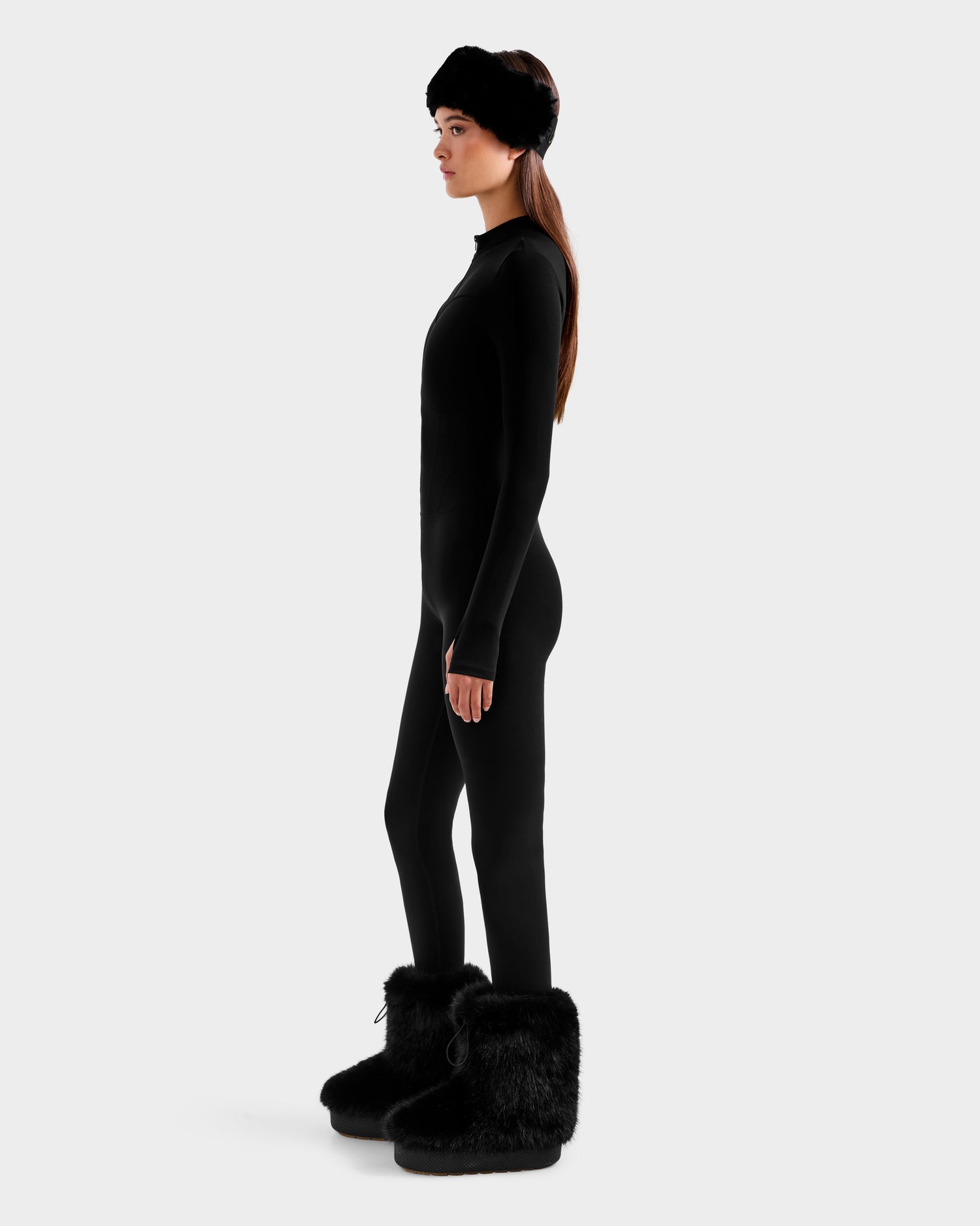 A woman stands in profile wearing the Rudsak IVY - 8524599 Black stretch bodysuit, paired with black furry boots and a matching fur headband, exuding sleek ski-chic style against a plain light gray background.