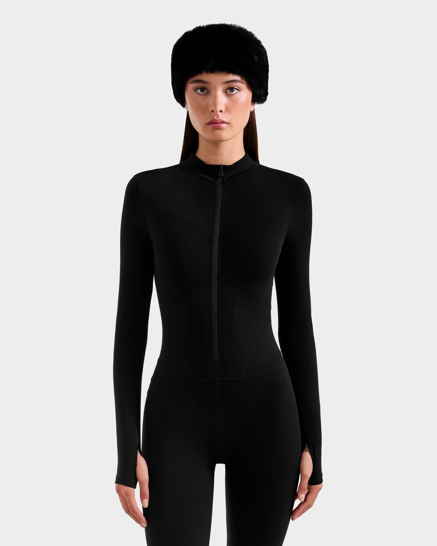 A woman stands facing forward in the IVY - 8524599 Black stretch bodysuit by Rudsak, featuring long sleeves and a front zipper. She wears a black fur hat and has long brown hair, showcasing ski-chic style against a light gray background.