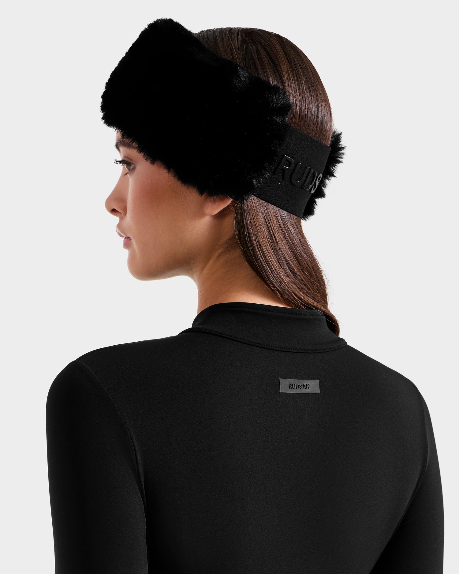 A woman with straight brown hair models the Rudsak IVY - 8524599 Black, a ski-chic zip-up bodysuit with a black fur headband and a small rectangular logo on the upper back, facing away against a plain light background.