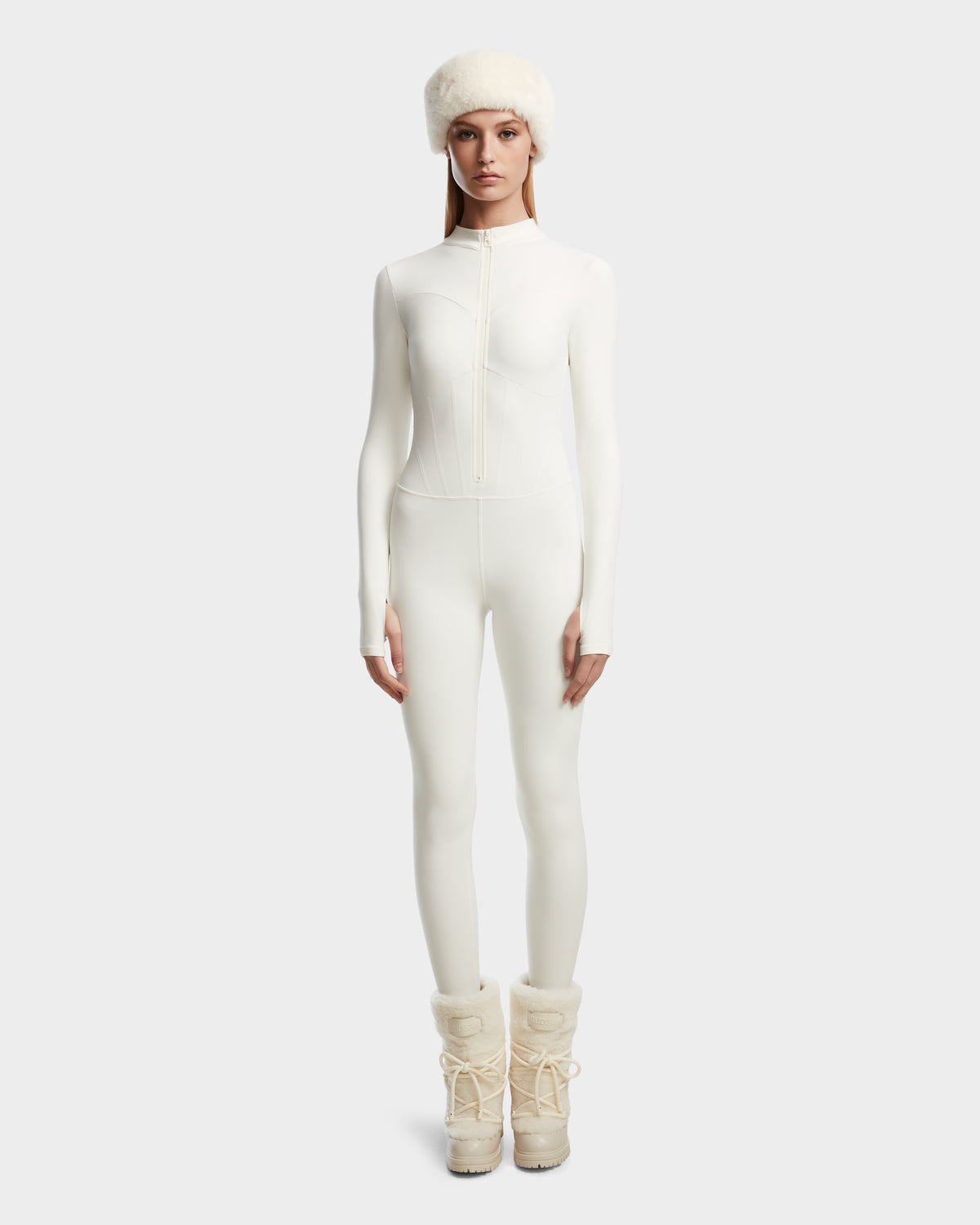 A woman stands facing forward, wearing Rudsaks IVY - 8524599 Iceland fitted white zip-up bodysuit, a fluffy white hat, and beige fur-lined winter boots against a plain light background.