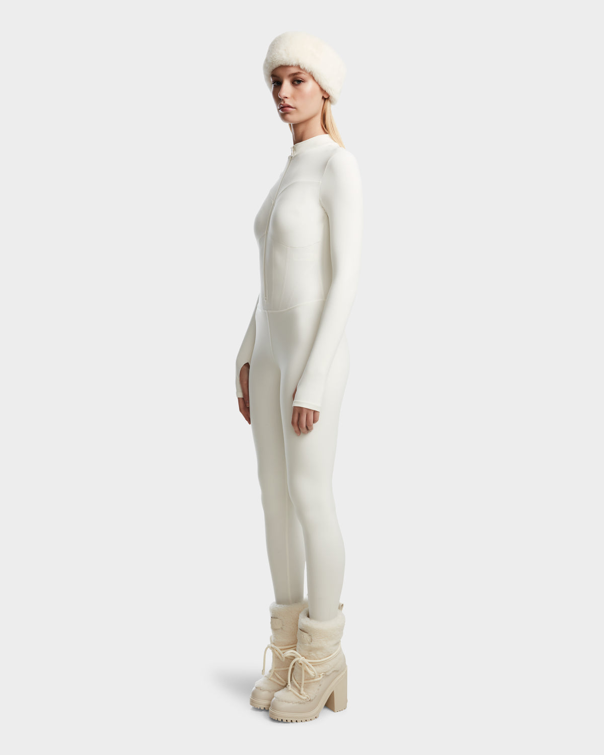 A woman poses in profile in Rudsaks IVY - 8524599 Iceland fitted stretch bodysuit, beige heeled winter boots, and a fluffy white hat, showcasing ski-chic style against a plain light background.