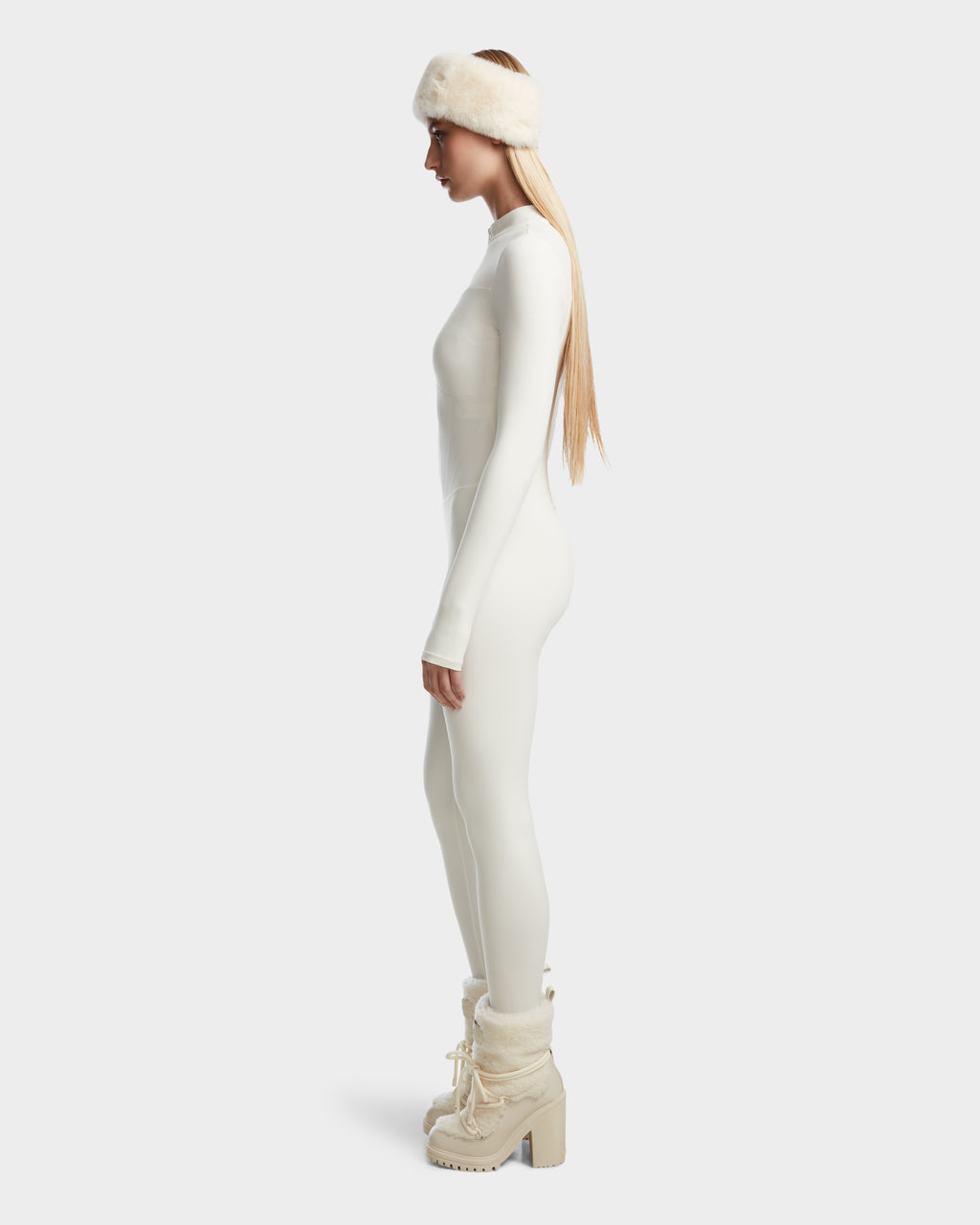 A woman in profile wears the Rudsak IVY - 8524599 Iceland fitted stretch bodysuit, a fur headband, long blonde hair, and white fur-lined chunky heeled boots against a plain light background.