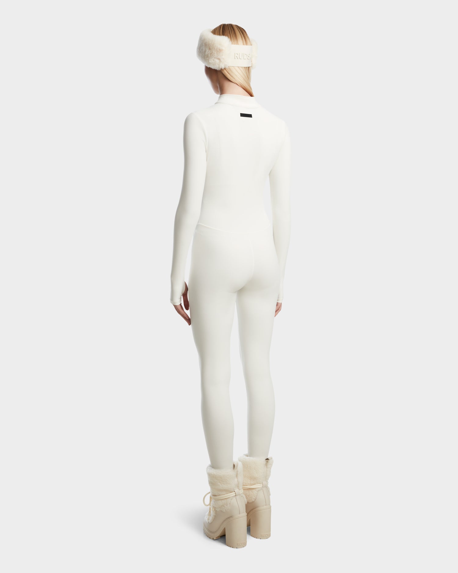 A person models the Rudsak IVY - 8524599 Iceland womens stretch bodysuit with cream fur earmuffs and beige lace-up chunky-heeled winter boots, showcasing a ski-chic style against a plain light background.