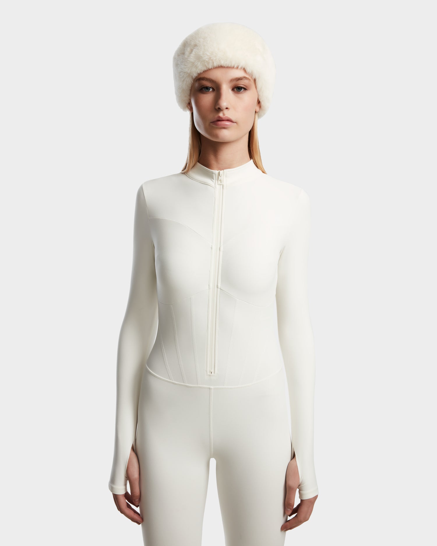 A woman stands facing forward in the Rudsak IVY - 8524599 Iceland fitted, long-sleeve white zip-up bodysuit with a front zipper and a fluffy white hat, showcasing ski-chic style against a plain light background.