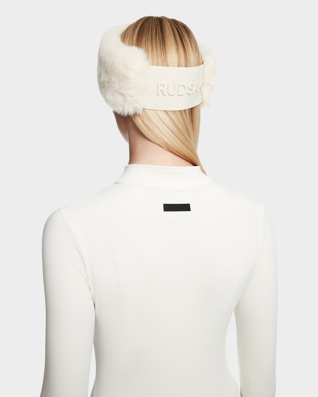 A person with straight blonde hair wears Rudsak IVY - 8524599 Iceland white furry earmuffs and a white high-collared zip-up bodysuit, shown from the back, exuding a sleek ski-chic vibe with RUDSAK displayed on the band.