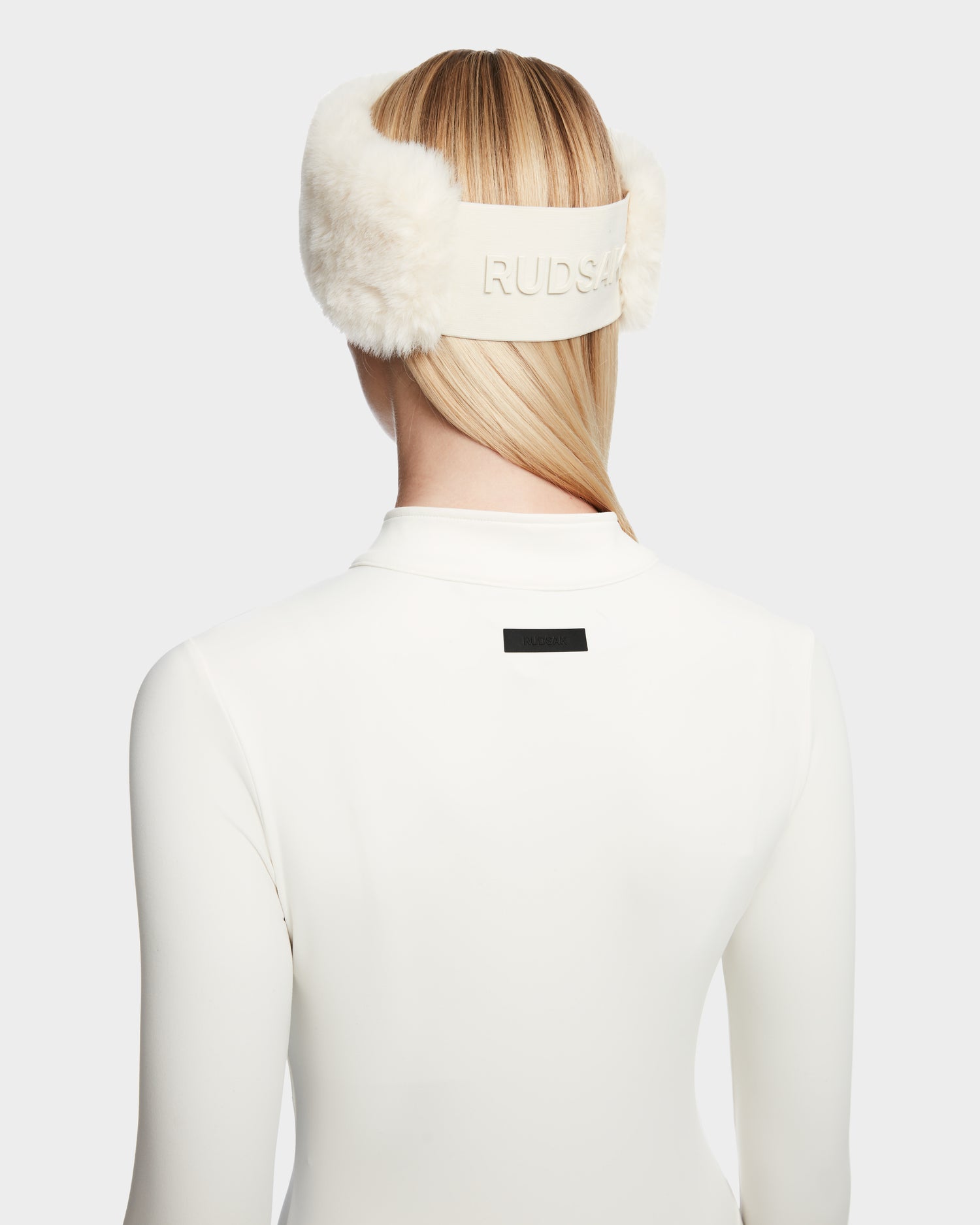 A person with straight blonde hair wears Rudsak IVY - 8524599 Iceland white furry earmuffs and a white high-collared zip-up bodysuit, shown from the back, exuding a sleek ski-chic vibe with RUDSAK displayed on the band.
