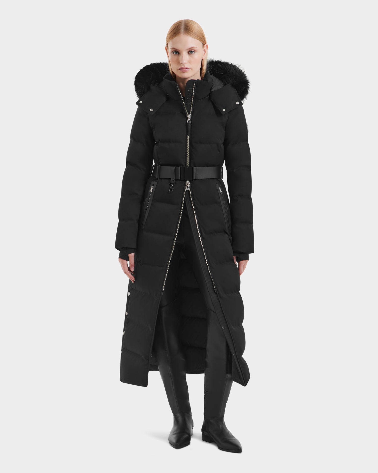 A woman stands against a plain white background wearing the Rudsak IZABELLA H - 8125644 Black puffer coat with RDS-certified down, a removable faux fur-trimmed hood, and wide belt, styled with knee-high boots.