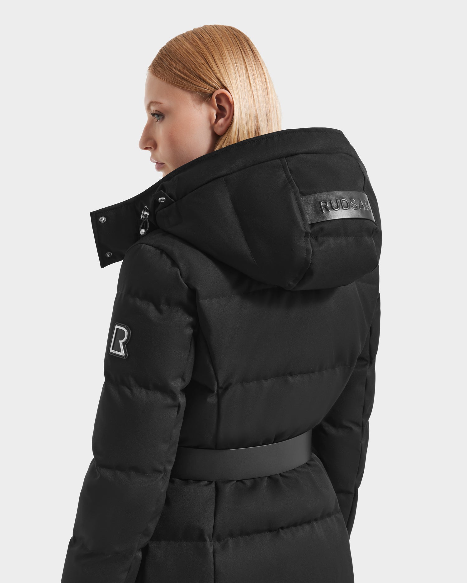 A woman with straight blonde hair wears the Rudsak IZABELLA H - 8125644 Black puffer coat, featuring a belt, shiny RUDSAK logo on the hood, R patch on the sleeve, and warm RDS-certified down. Shown from back and side for comfort and style.