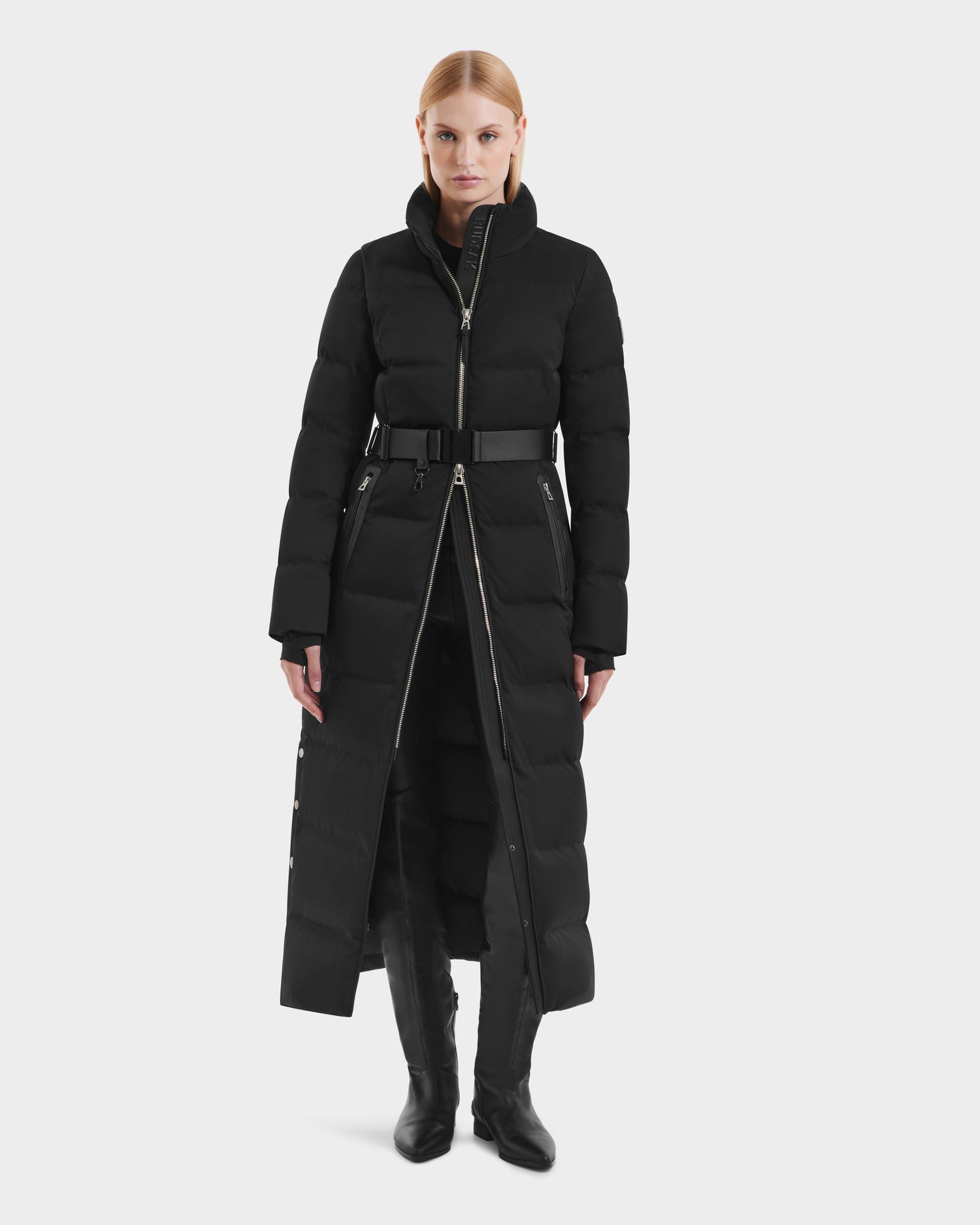 A woman faces forward in the Rudsak IZABELLA H - 8125644 Black women’s puffer coat with RDS-certified down, high collar, belted waist, front zippers, and black boots against a plain white background.
