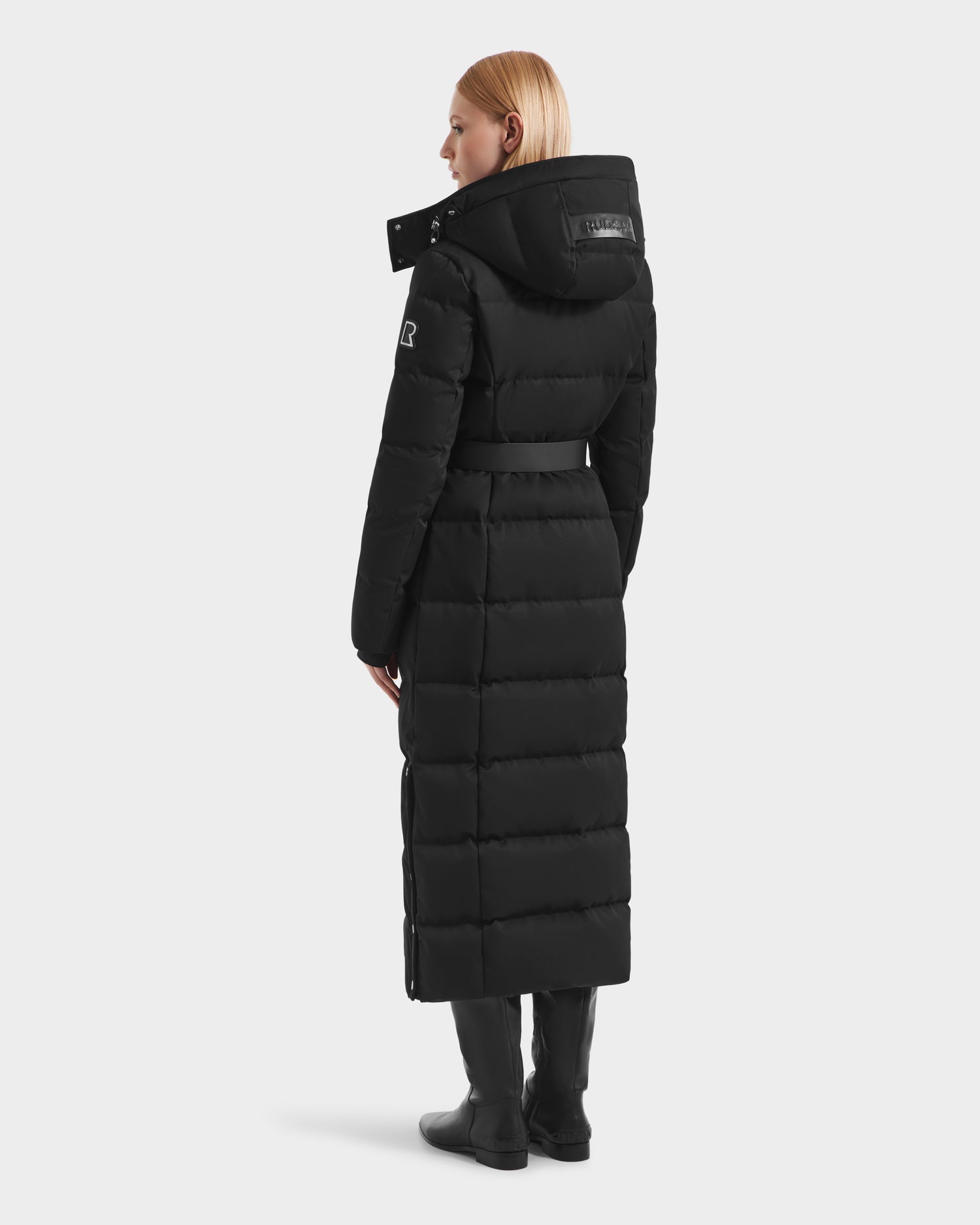 A woman with straight blonde hair wears the Rudsak IZABELLA H - 8125644 Black long puffer coat with a hood and belt, paired with black boots. She stands facing away, looking slightly to the side against a plain white background.