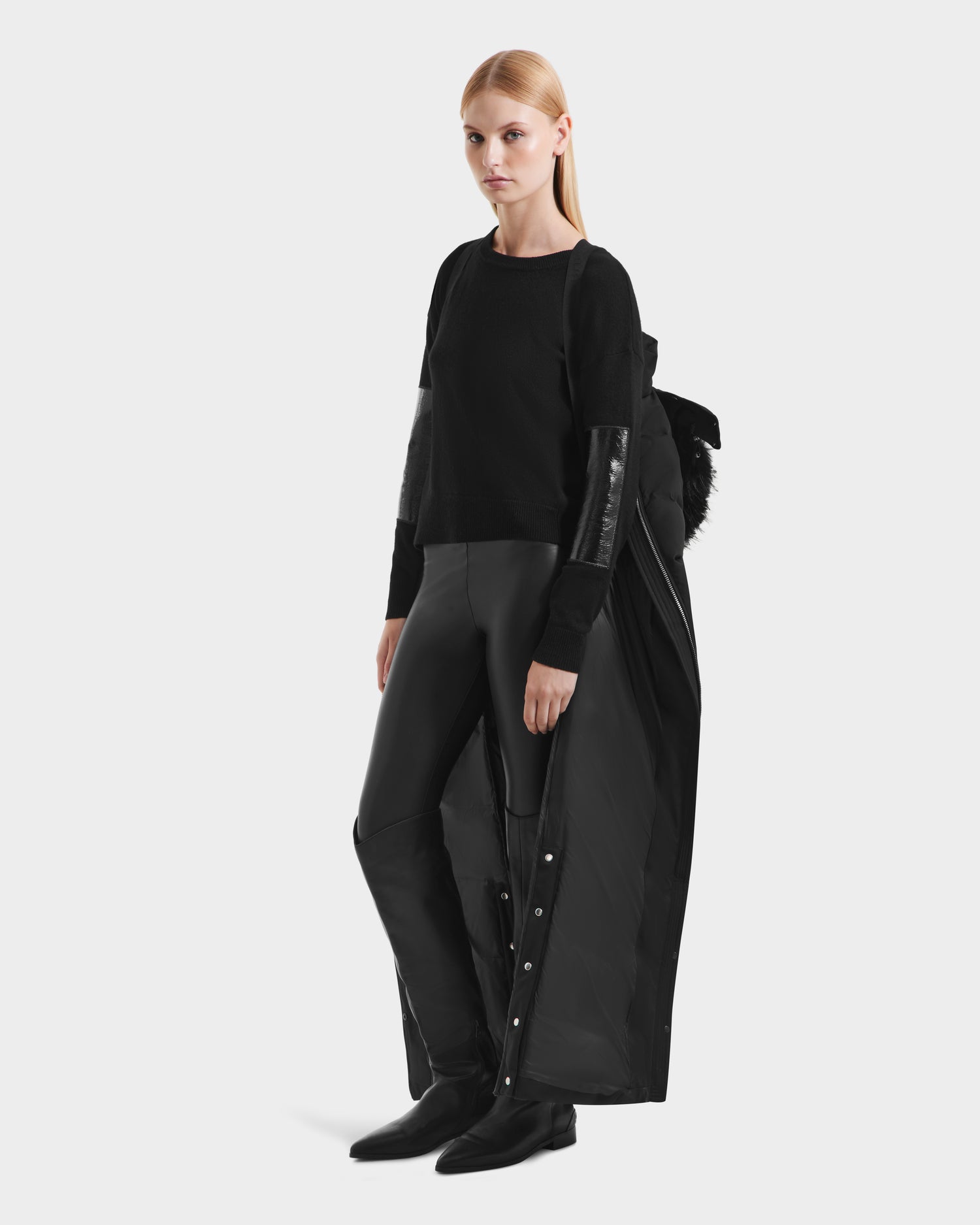 A woman with straight blonde hair wears the Rudsak IZABELLA H - 8125644 Black women’s puffer coat with removable faux fur draped over her shoulders, styled with black boots and fitted pants against a plain white background.