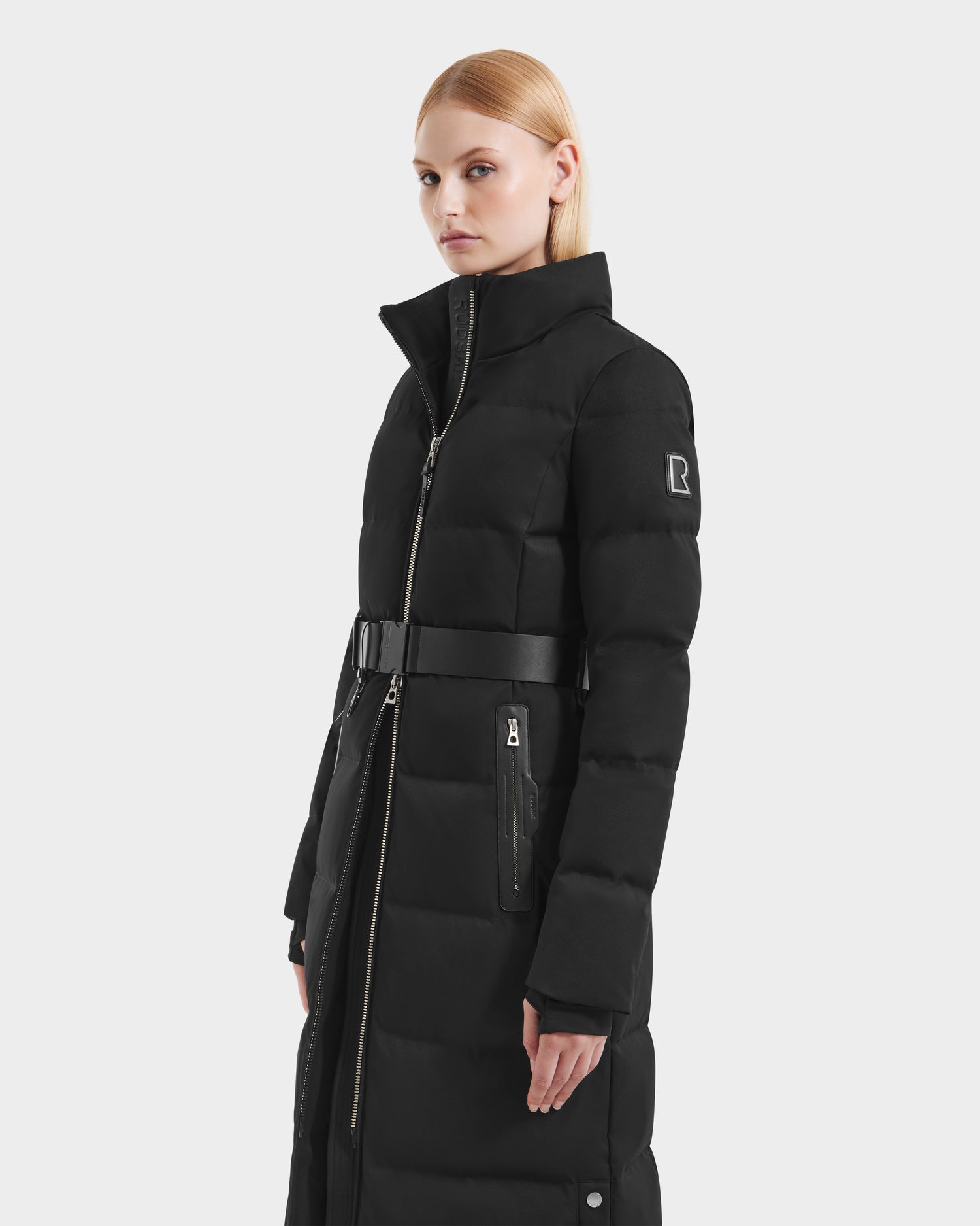 A woman with straight blonde hair models the Rudsak IZABELLA H - 8125644 Black women’s puffer coat featuring a high collar, belt, zippered pockets, and RDS-certified down, standing against a plain white background.