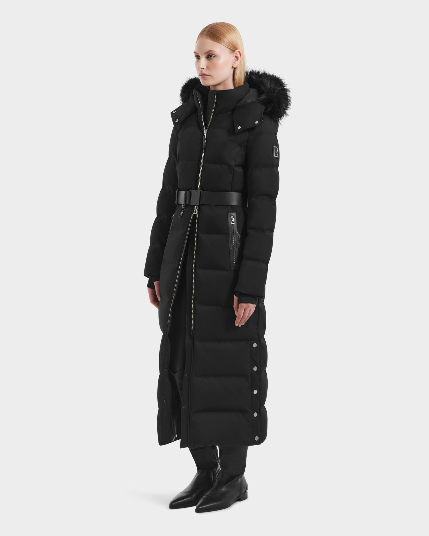 A woman stands slightly to the side wearing the Rudsak IZABELLA H - 8125644 Black puffer coat with RDS-certified down, a removable faux fur-trimmed hood, belted waist, and side snap buttons. She pairs it with black boots and has straight blonde hair.