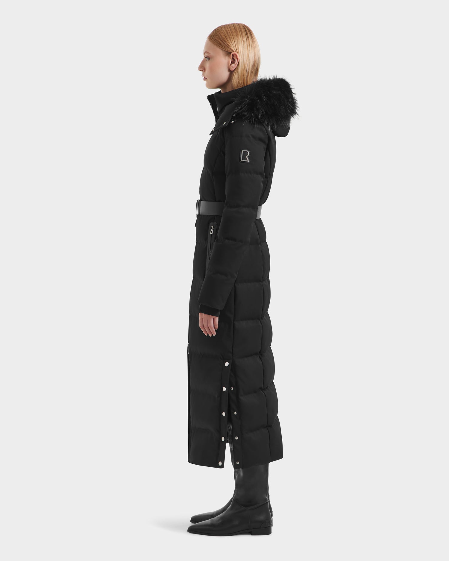 A woman with straight blonde hair models the Rudsak IZABELLA H - 8125644 Black women’s puffer coat, featuring a removable faux fur-trimmed hood and side snap buttons, paired with knee-high black boots against a plain light background.
