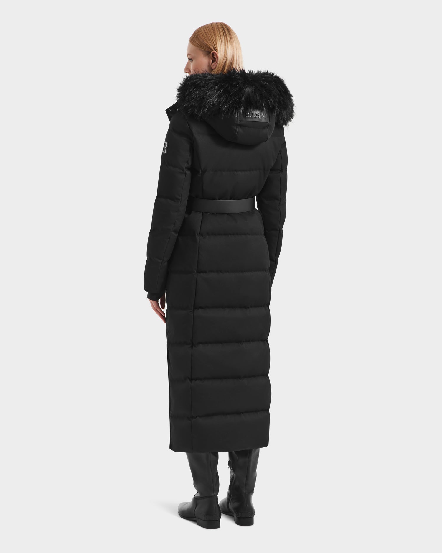 A blonde woman, seen from behind, wears the Rudsak IZABELLA H - 8125644 Black women’s puffer coat with RDS-certified down, a removable faux fur-trimmed hood, waist belt, and black boots against a plain light background.