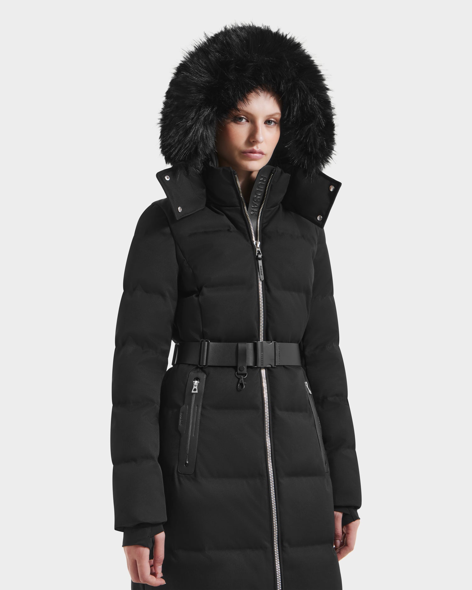 A person stands against a plain white background wearing the Rudsak IZABELLA H - 8125644 Black, a long women’s puffer coat with a belt and a large, removable faux fur-trimmed hood.
