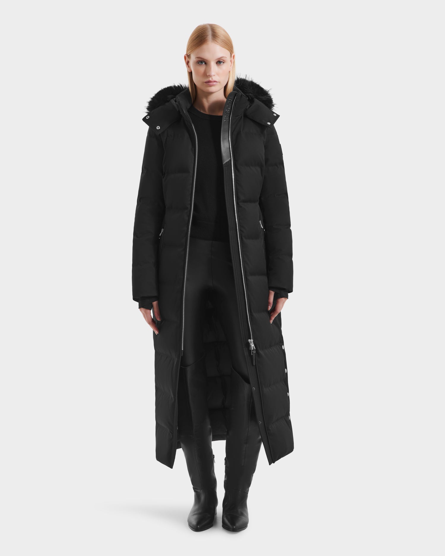 A woman stands facing forward against a white background, wearing the Rudsak IZABELLA H - 8125644 Black women's long puffer coat with a hood, paired with a black sweater, pants, and boots.