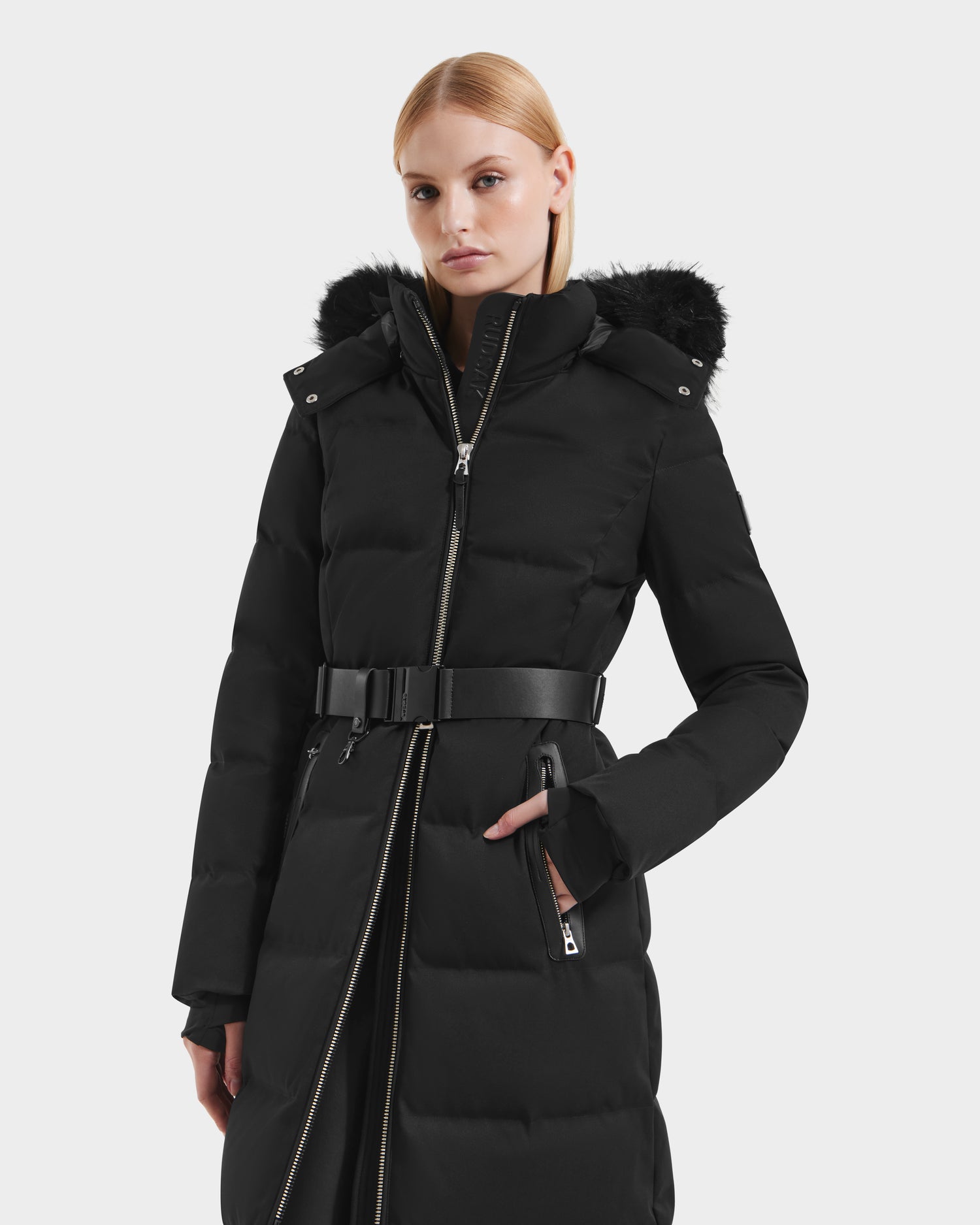 A woman wears the Rudsak IZABELLA H - 8125644 Black puffer winter coat with a belted waist, zippered front, and black faux fur-trimmed hood, standing against a plain light background.