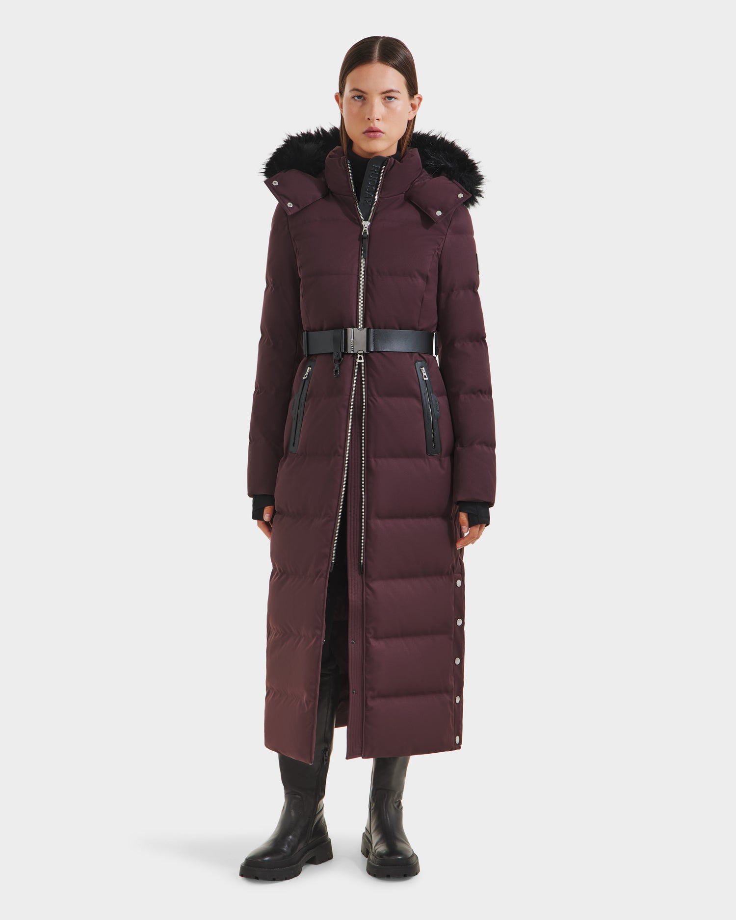 A woman faces forward, wearing the Rudsak IZABELLA H - 8125644 Bordeaux—a long maroon puffer with a black fur-trimmed hood and belt—paired with gloves and boots against a plain white background.