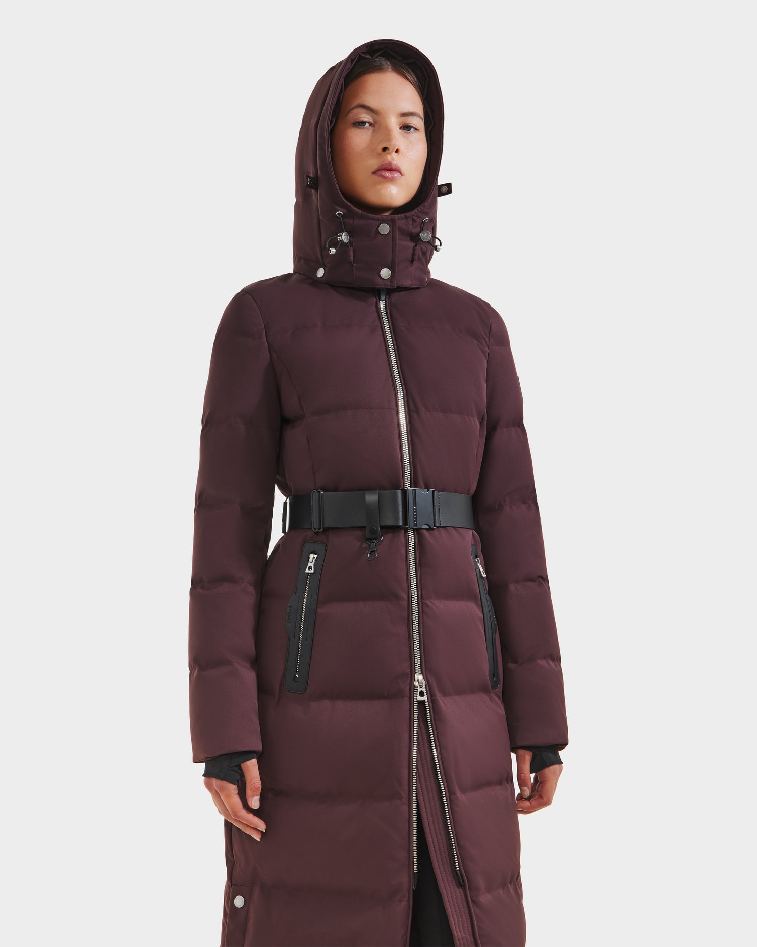 A woman models the Rudsak IZABELLA H - 8125644 Bordeaux puffer coat with a black belt and zipper details, set against a plain white background.