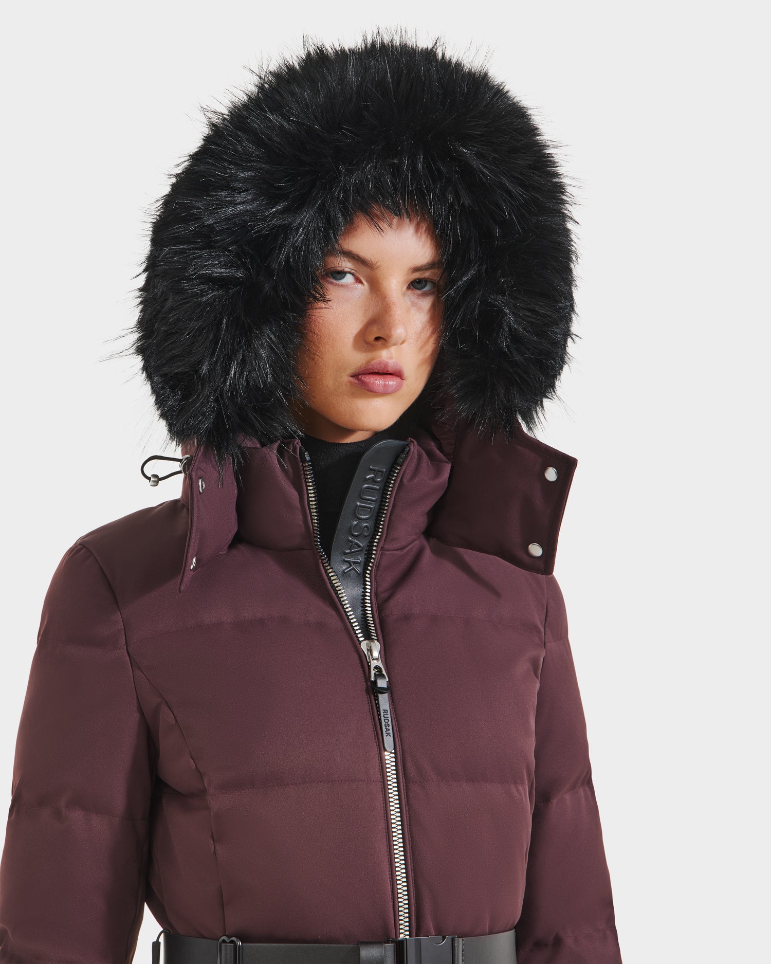 A woman models the Rudsak IZABELLA H - 8125644 Bordeaux puffer jacket, featuring a large black faux fur-trimmed hood and black zipper, against a plain white background.