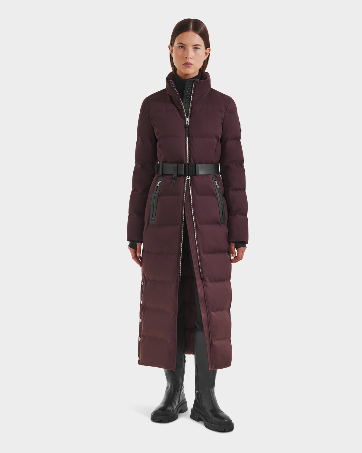 A woman faces forward wearing the Rudsak IZABELLA H - 8125644 Bordeaux, a long belted dark burgundy puffer with a high collar and zippered pockets, styled with a black turtleneck, black pants, and chunky boots against a white background.