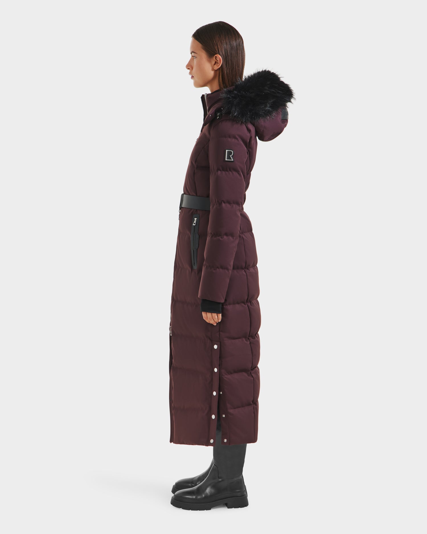 A woman stands in profile wearing the Rudsak IZABELLA H - 8125644 Bordeaux, a long, dark maroon puffer coat with a black fur-trimmed hood, black belt, black boots, and button details along the side.