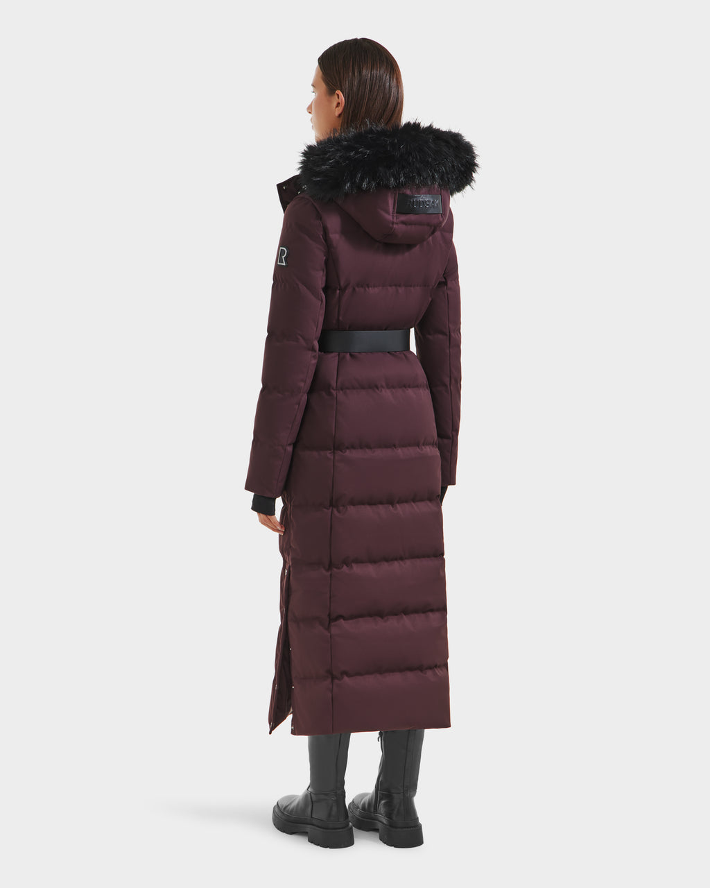 A woman faces away, wearing the Rudsak IZABELLA H - 8125644 Bordeaux puffer with black fur-trimmed hood, belted waist, and black boots. This long cold-weather coat extends below her knees for added warmth.
