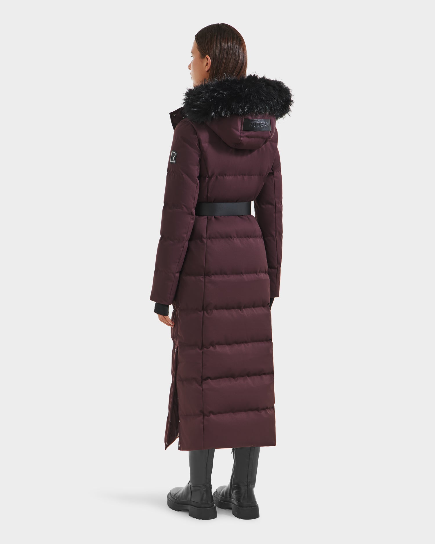 A woman faces away, wearing the Rudsak IZABELLA H - 8125644 Bordeaux puffer with black fur-trimmed hood, belted waist, and black boots. This long cold-weather coat extends below her knees for added warmth.