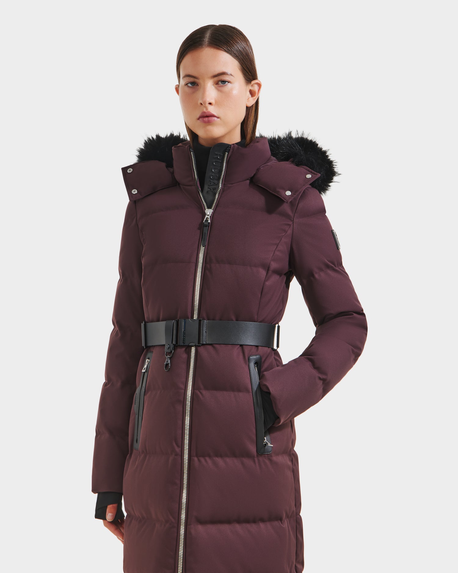 A woman stands facing forward against a plain white background, wearing the Rudsak IZABELLA H - 8125644 Bordeaux long puffer coat with a black belt, faux fur-trimmed hood, and black accents.
