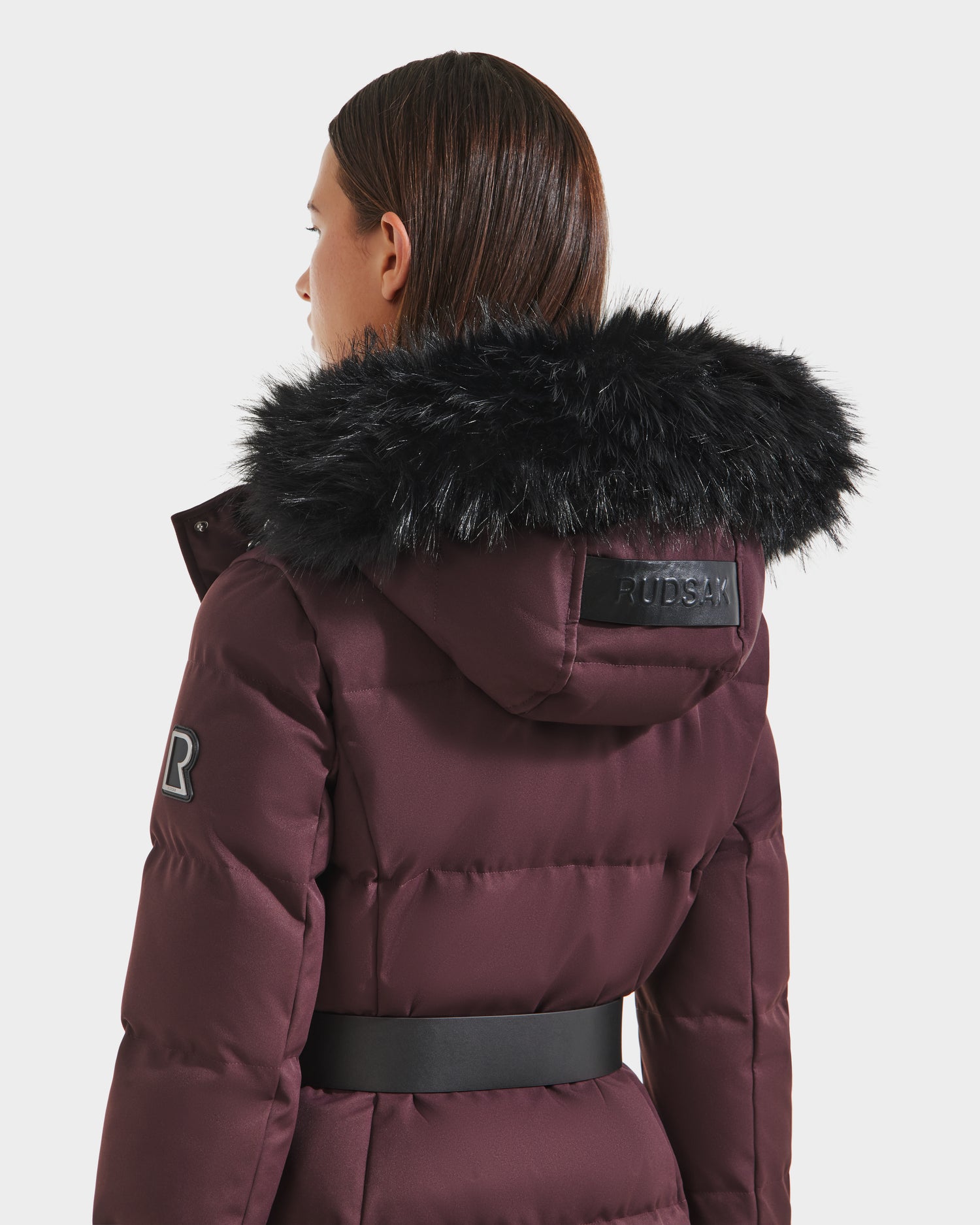 A person with straight brown hair is seen from the back wearing the Rudsak IZABELLA H - 8125644 Bordeaux women’s cold-weather puffer coat with RDS-certified down, black faux fur hood, R patch on the sleeve, and a wide black waist belt.